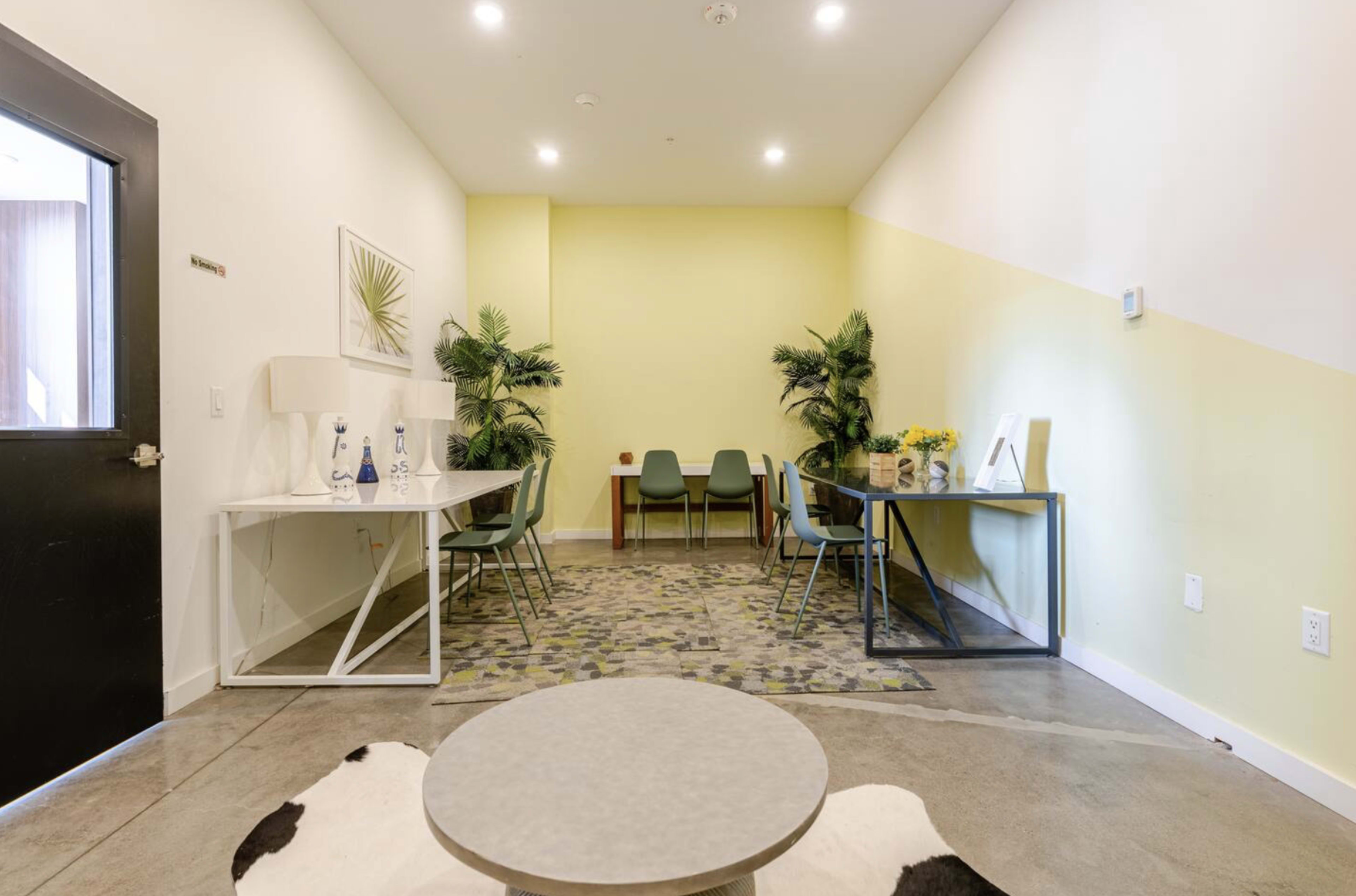 The image shows a modern room with a yellow accent wall, featuring a table and chairs, as well as a work desk, plants, and decorative elements on the floor.
