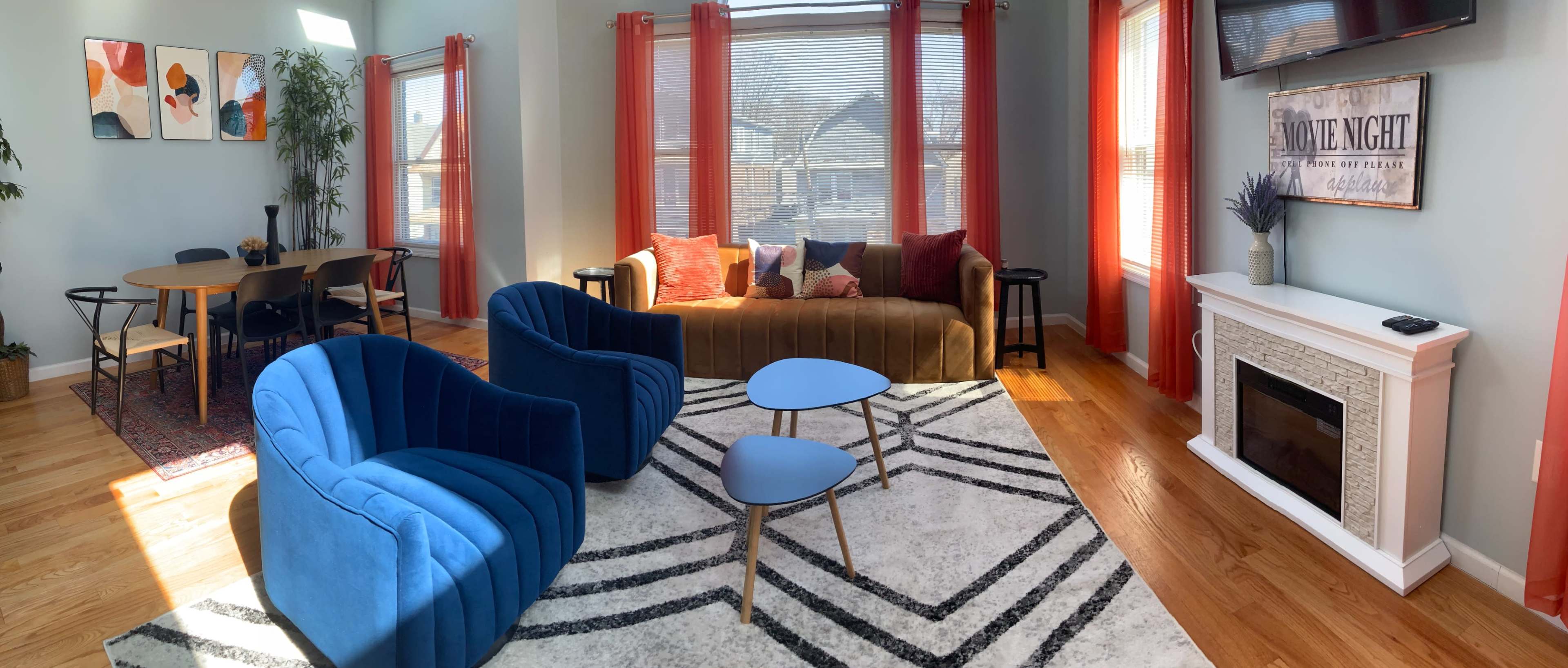 A modern living room with a blue sofa, two blue armchairs, a coffee table, a brown couch, and a dining area with a table and chairs, all featuring large windows and bright natural light.