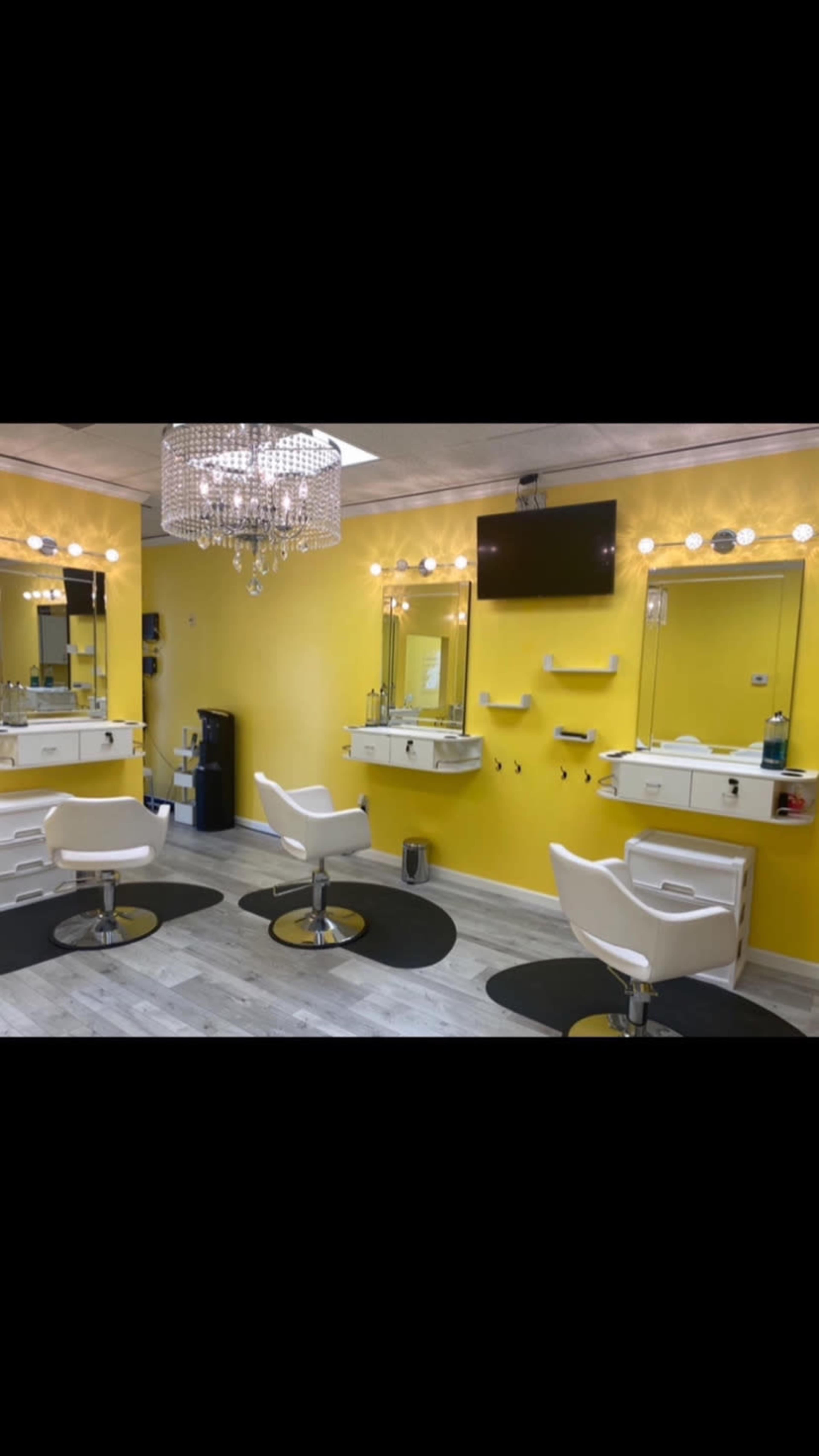 A brightly colored salon interior with two styling chairs, three mirrors, and a chandelier, all set against a yellow wall.