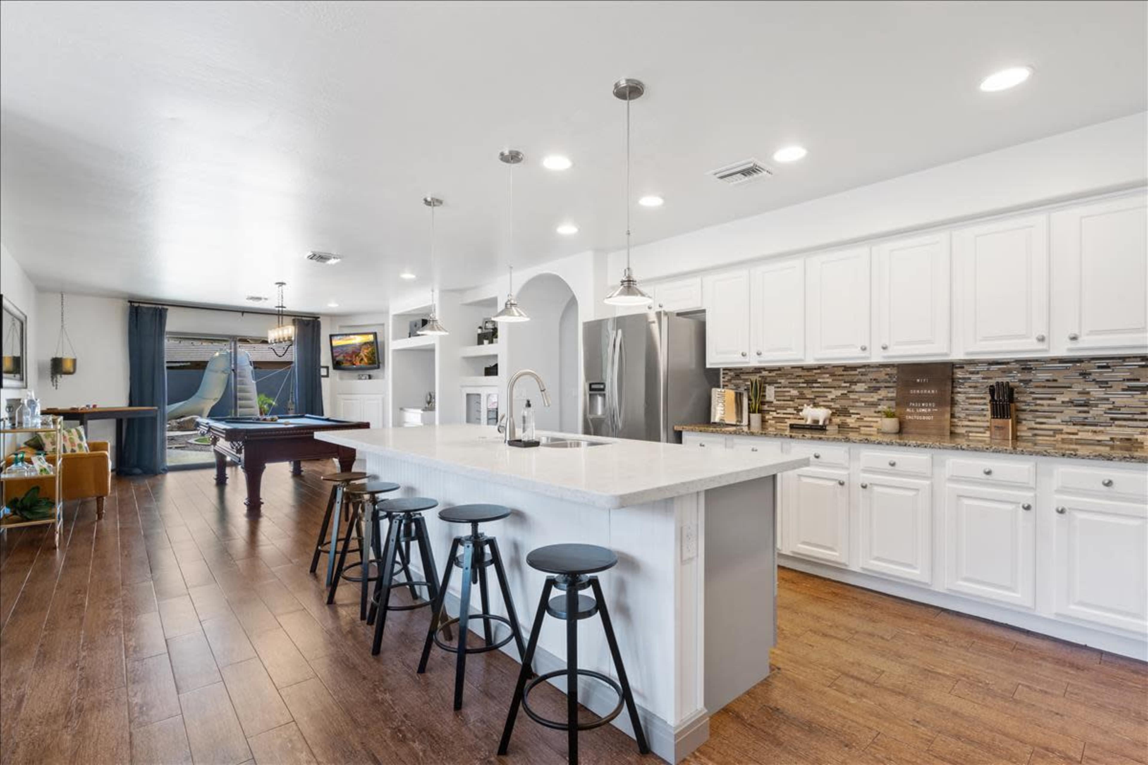 The image shows a modern kitchen with white cabinetry, a large island with bar stools, and a view into a spacious living area featuring a pool table.