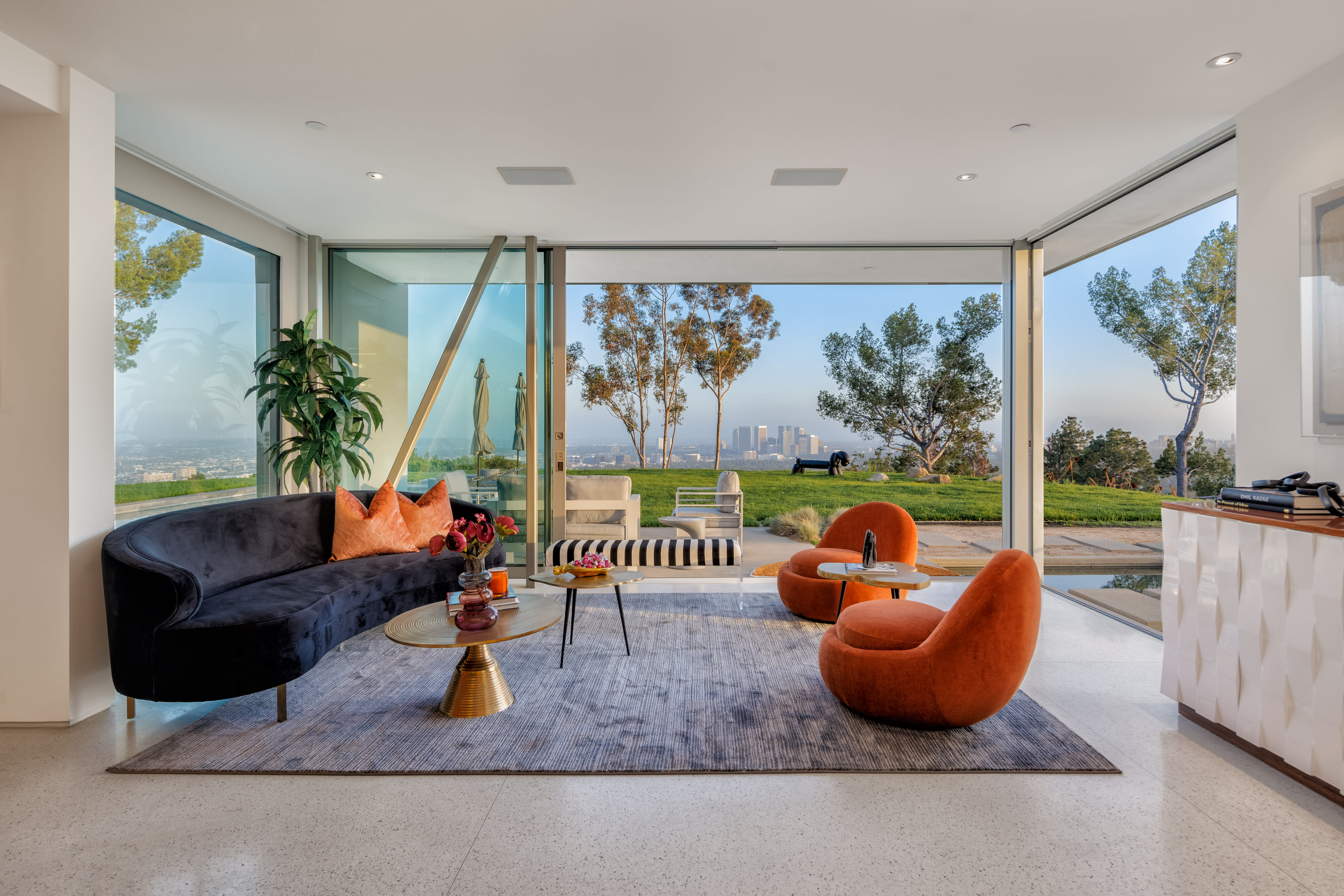 The modern living room features a large glass wall that opens to a view of the city, furnished with a curved black sofa, two orange chairs, and a glass coffee table on a large area rug.