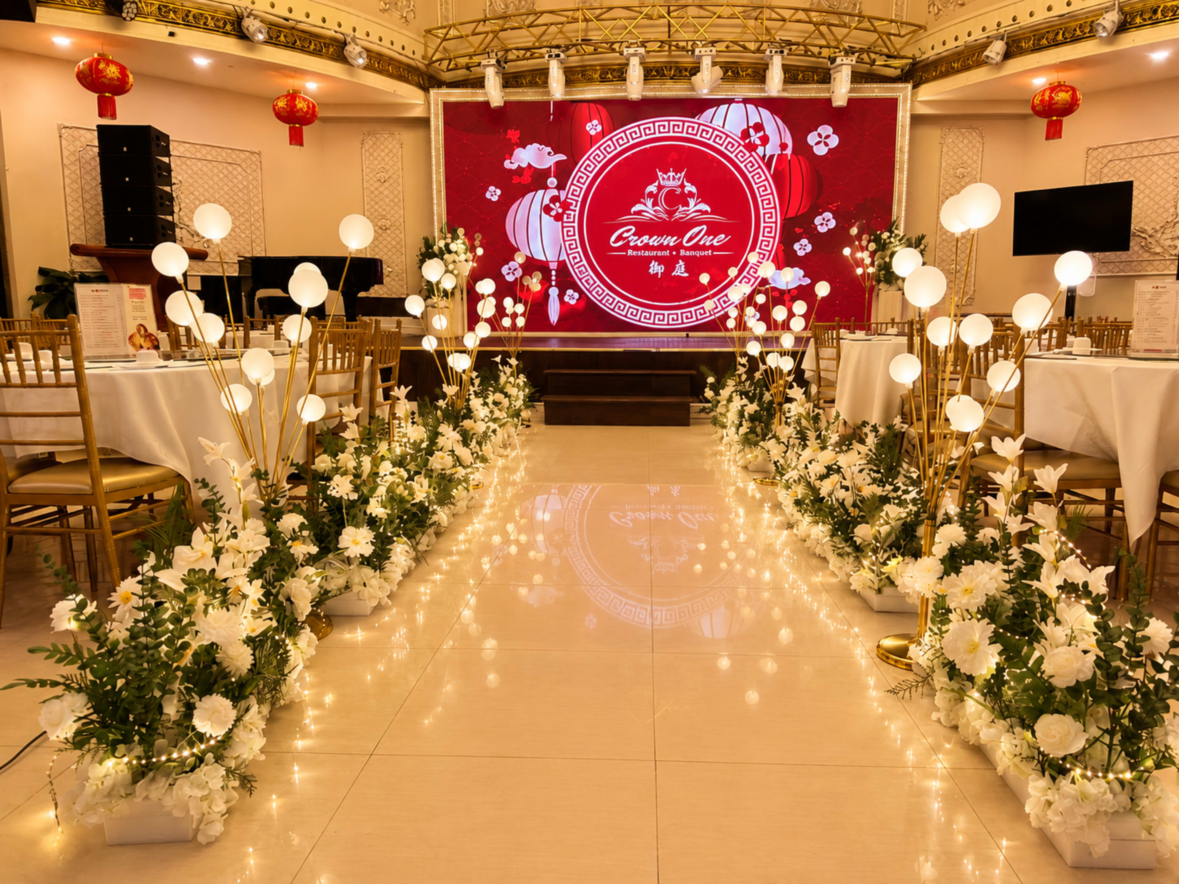 The image shows a decorated indoor event space with tables set for a celebration, flanked by illuminated floral arrangements and a large backdrop featuring a logo.