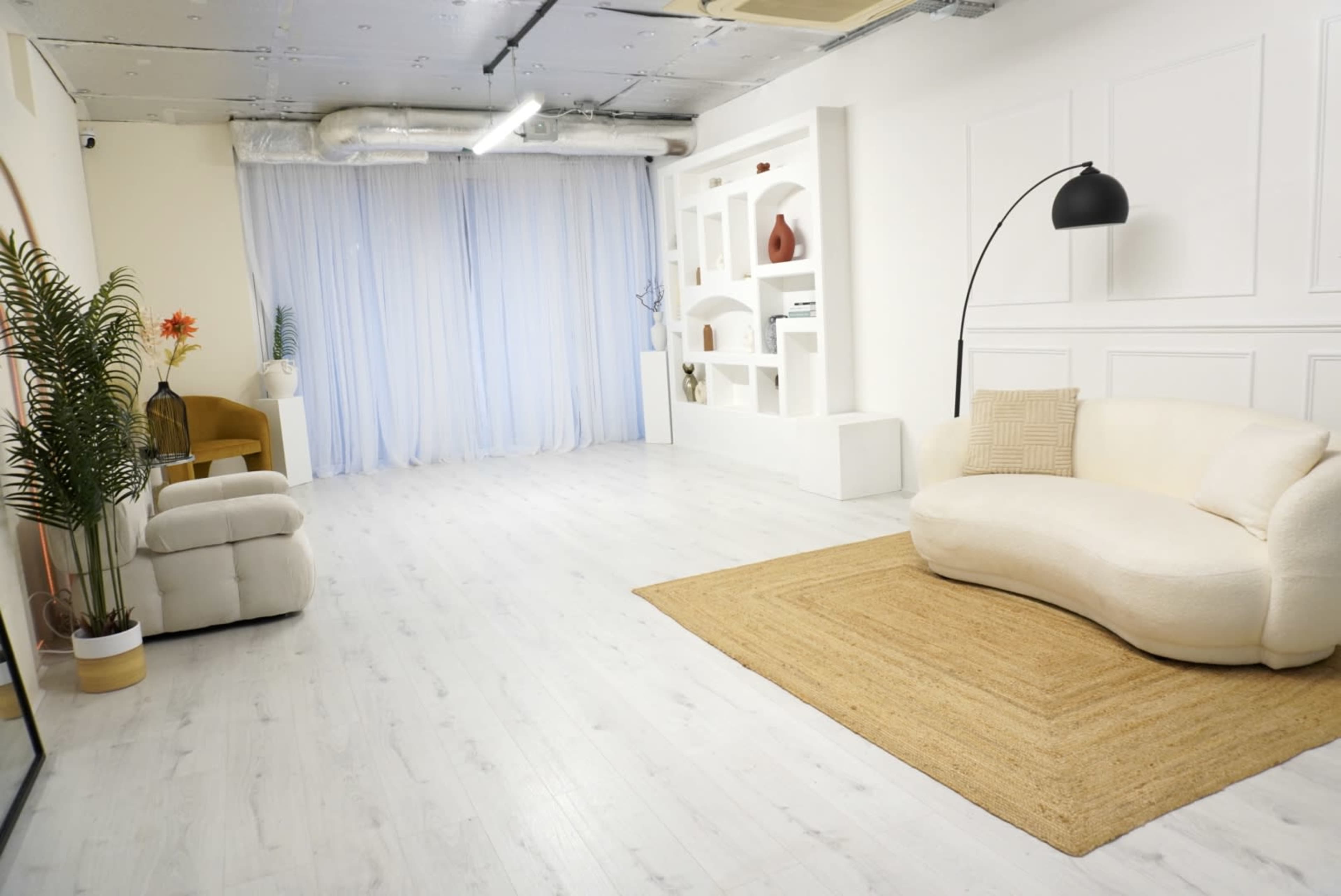 The image shows a bright, minimalistic room featuring a curved sofa, a chair, a jute rug, and a backdrop of sheer curtains along with decorative shelves on the wall.