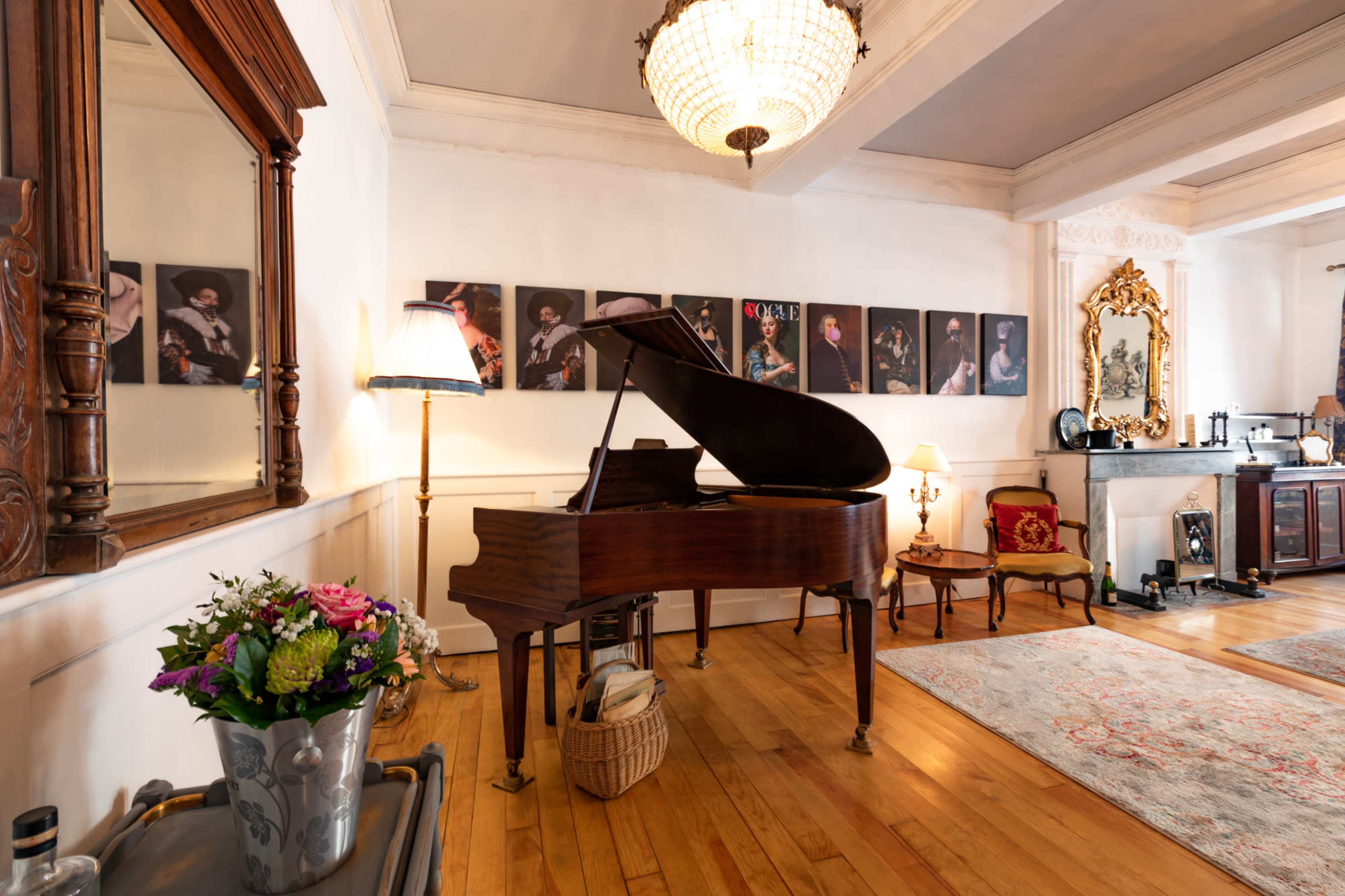 A grand piano is positioned in a room adorned with framed photographs, a large chandelier hangs overhead, and a rug covers the wooden floor.