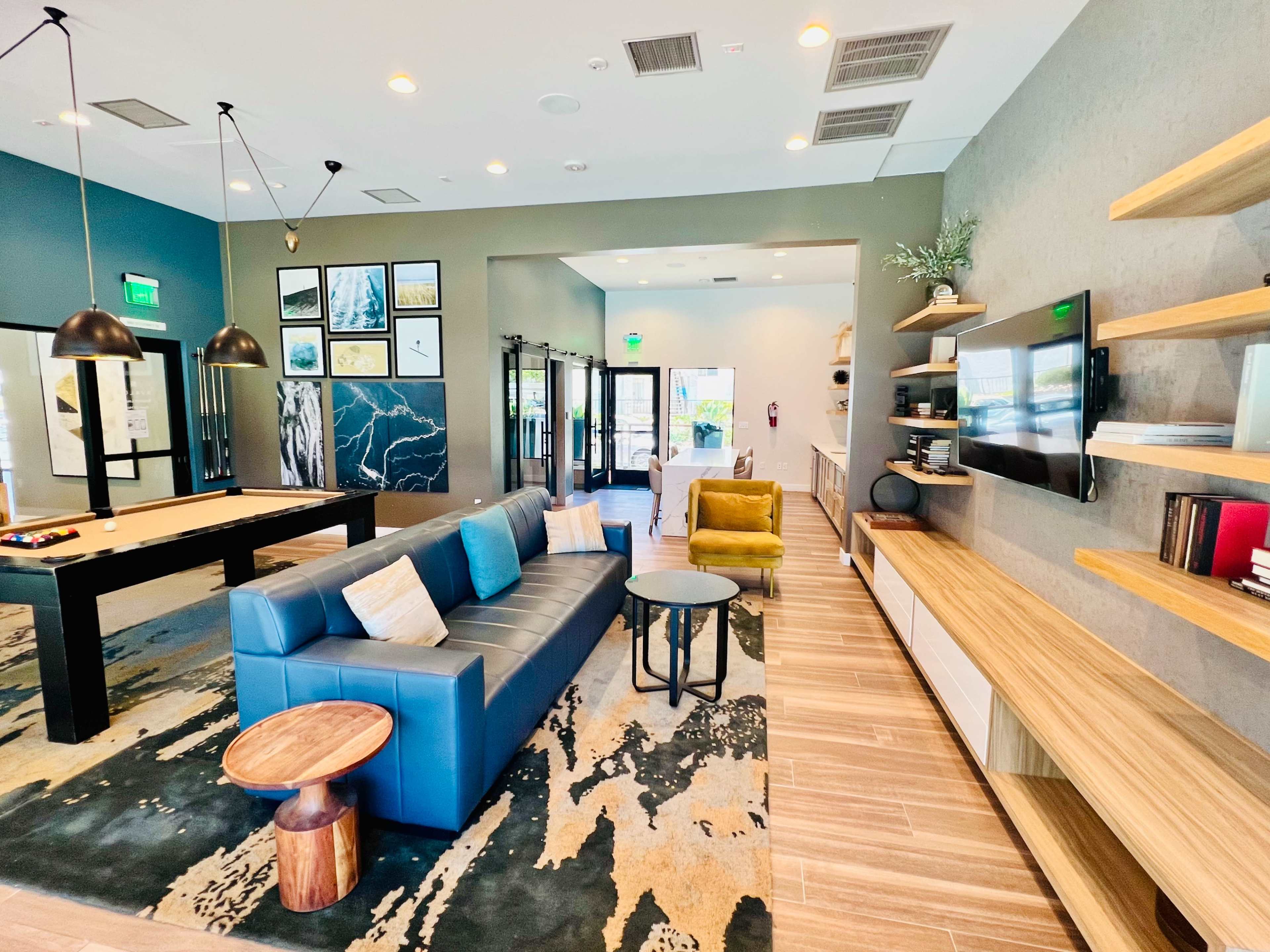 A modern lobby with a blue sofa, yellow chair, pool table, and a wall-mounted television surrounded by bookshelves.