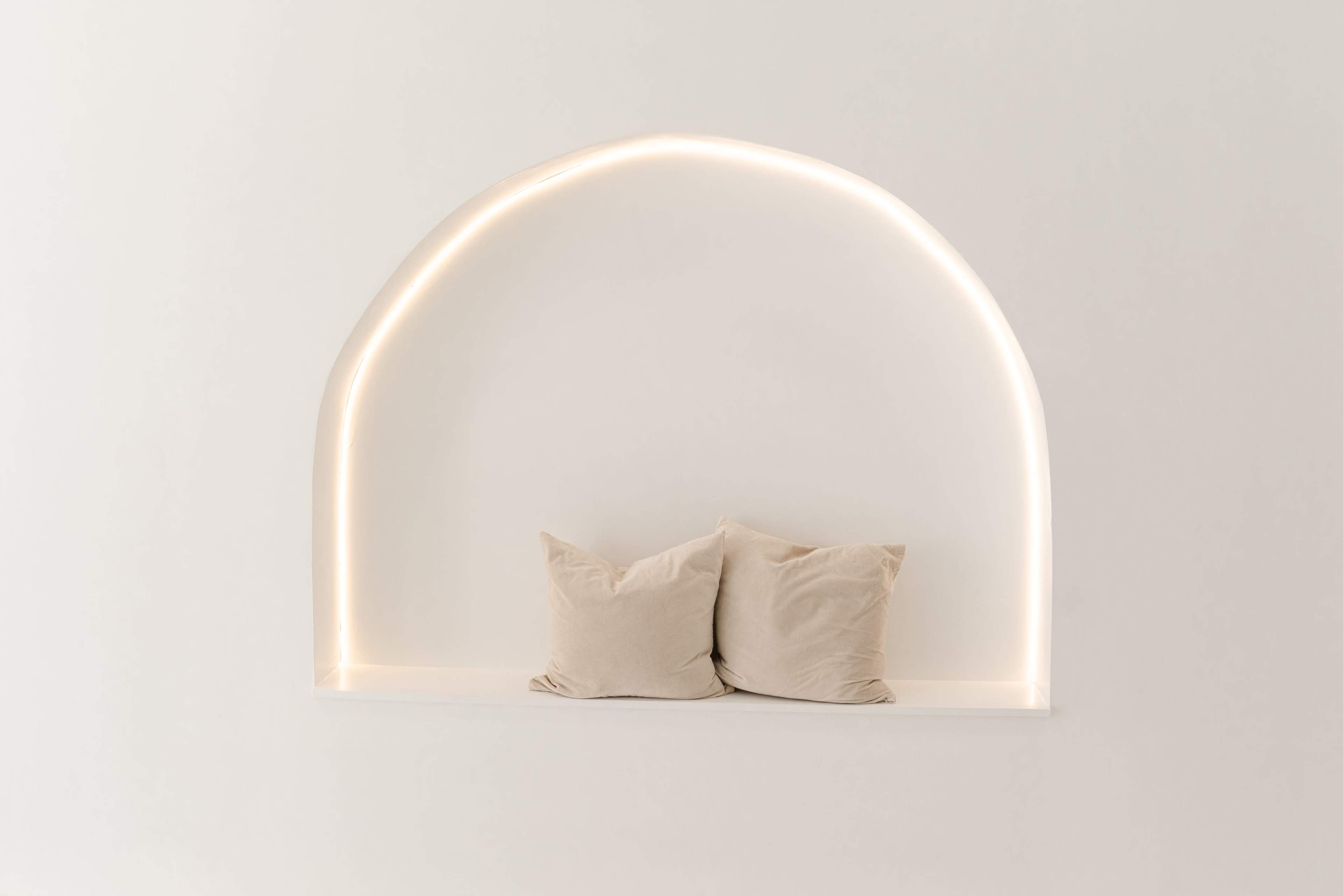 A minimalist wall niche features two plush pillows illuminated by a soft, curved light.