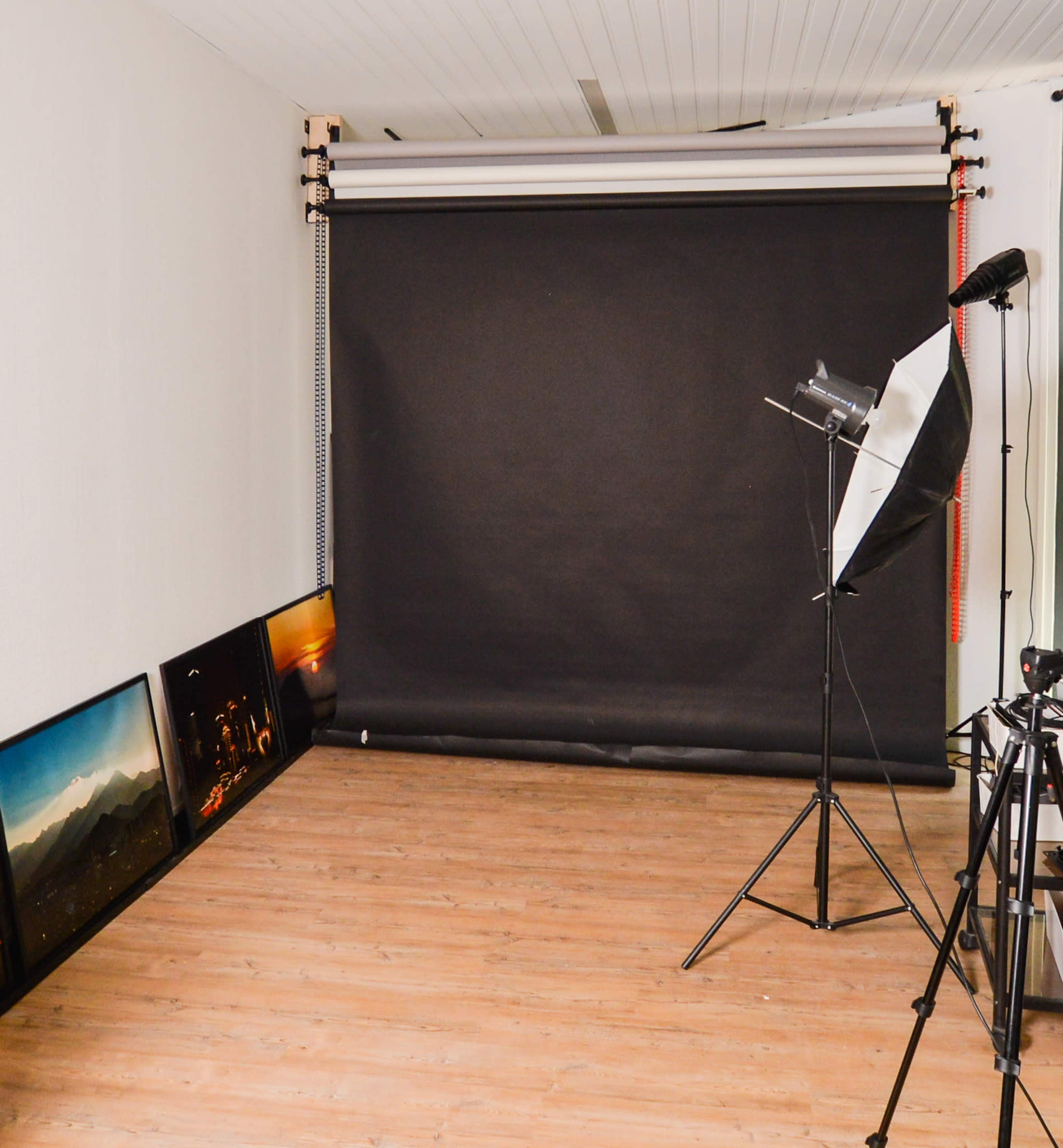 A photography studio with a black backdrop setup, a light source on a stand, and framed photos leaning against the wall.