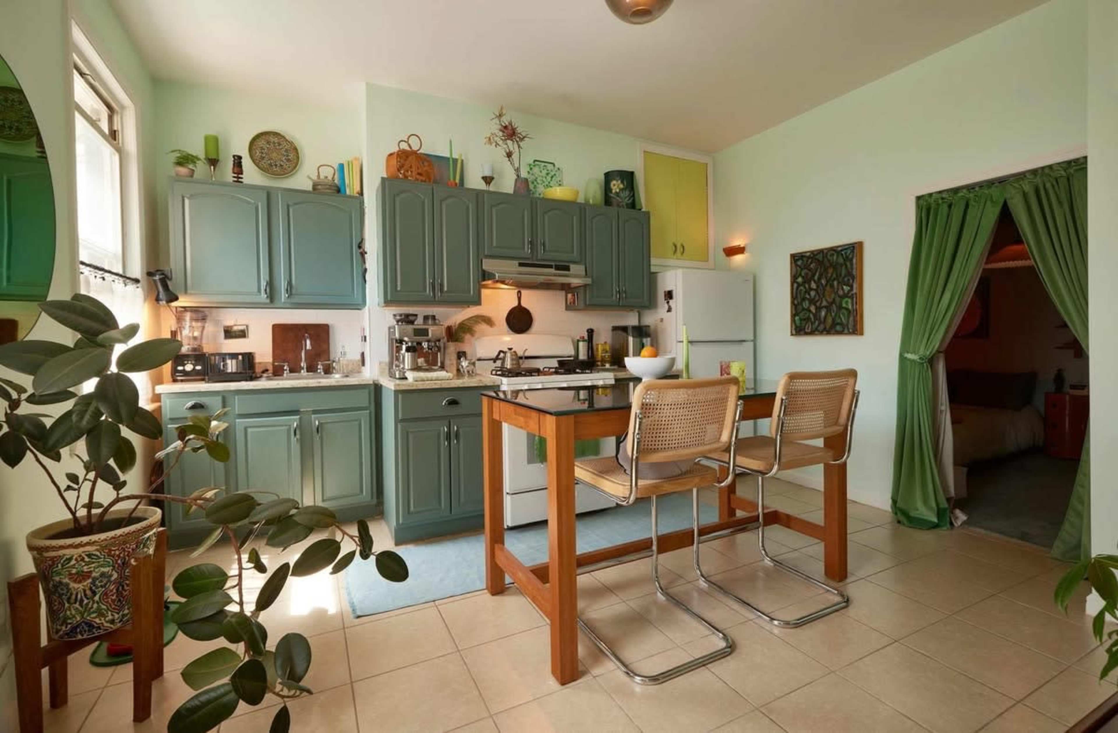 The kitchen features teal cabinets, a central wooden table with two bar stools, and various plants placed around the space.