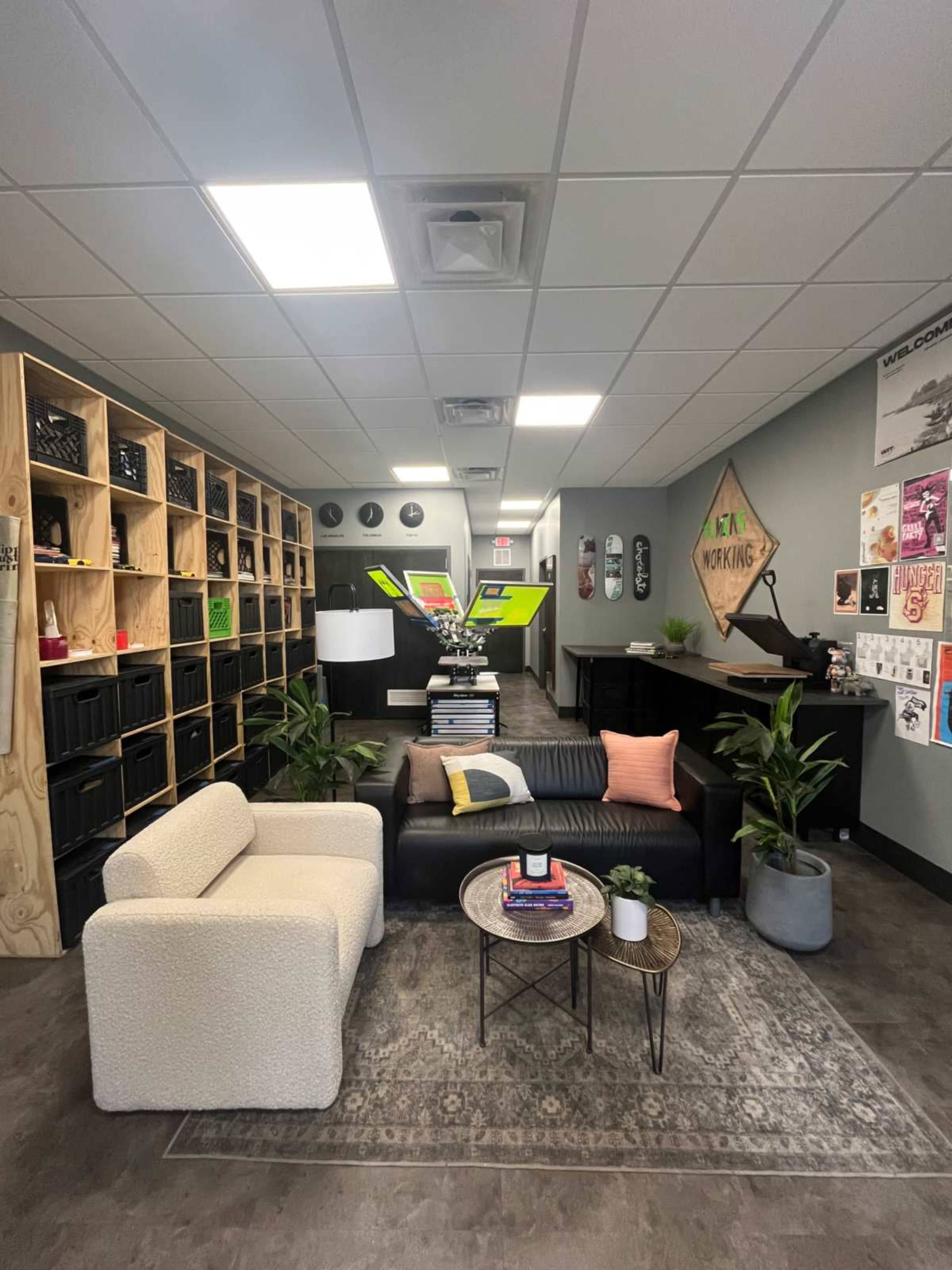 The image shows a modern office interior featuring a lounge area with two couches, decorative plants, and shelves filled with various items along a corridor with bright lighting.