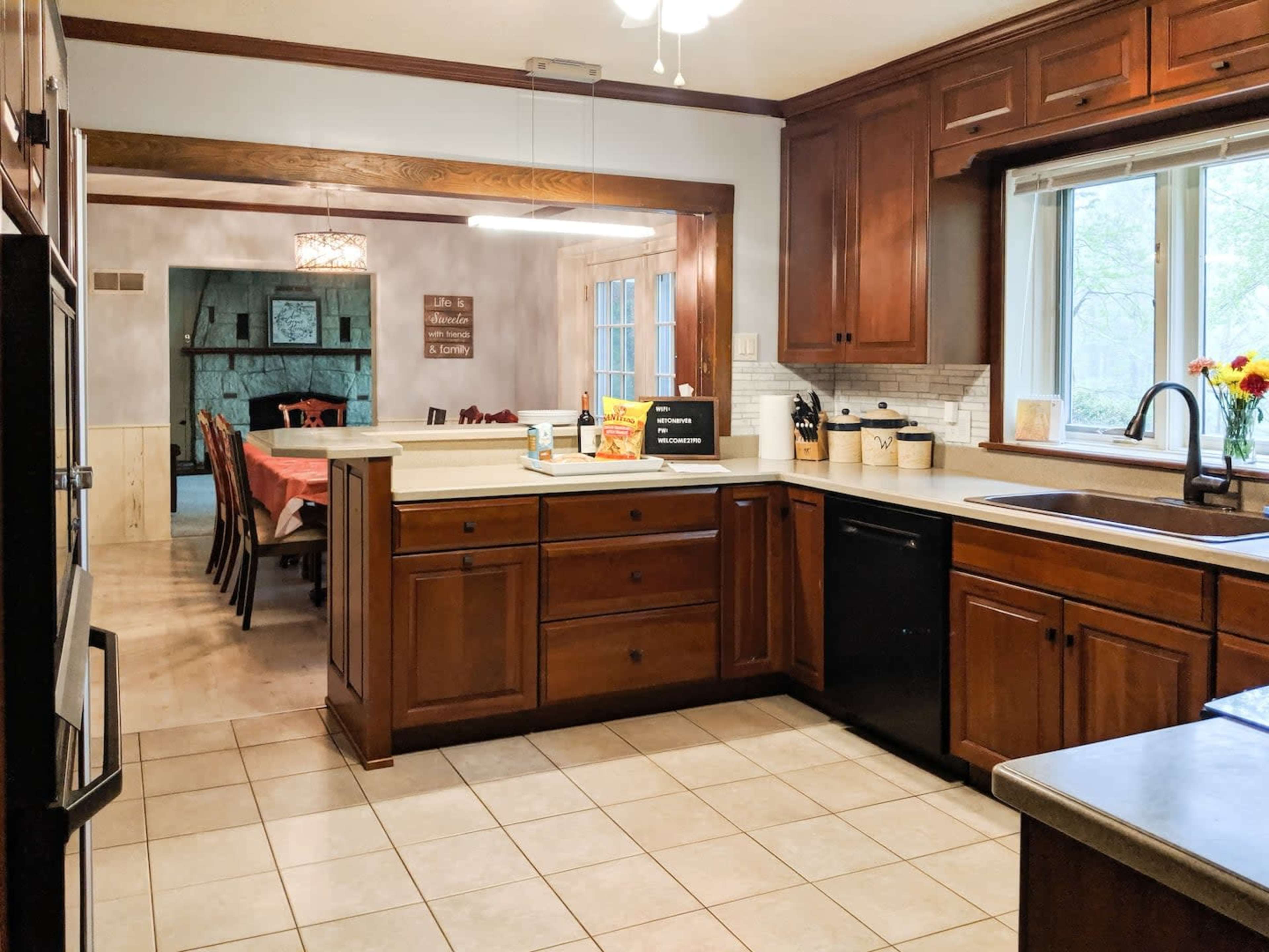 A kitchen with wooden cabinets, a sink, a black dishwasher, and a tiled floor, with an adjacent dining area visible through an open doorway.