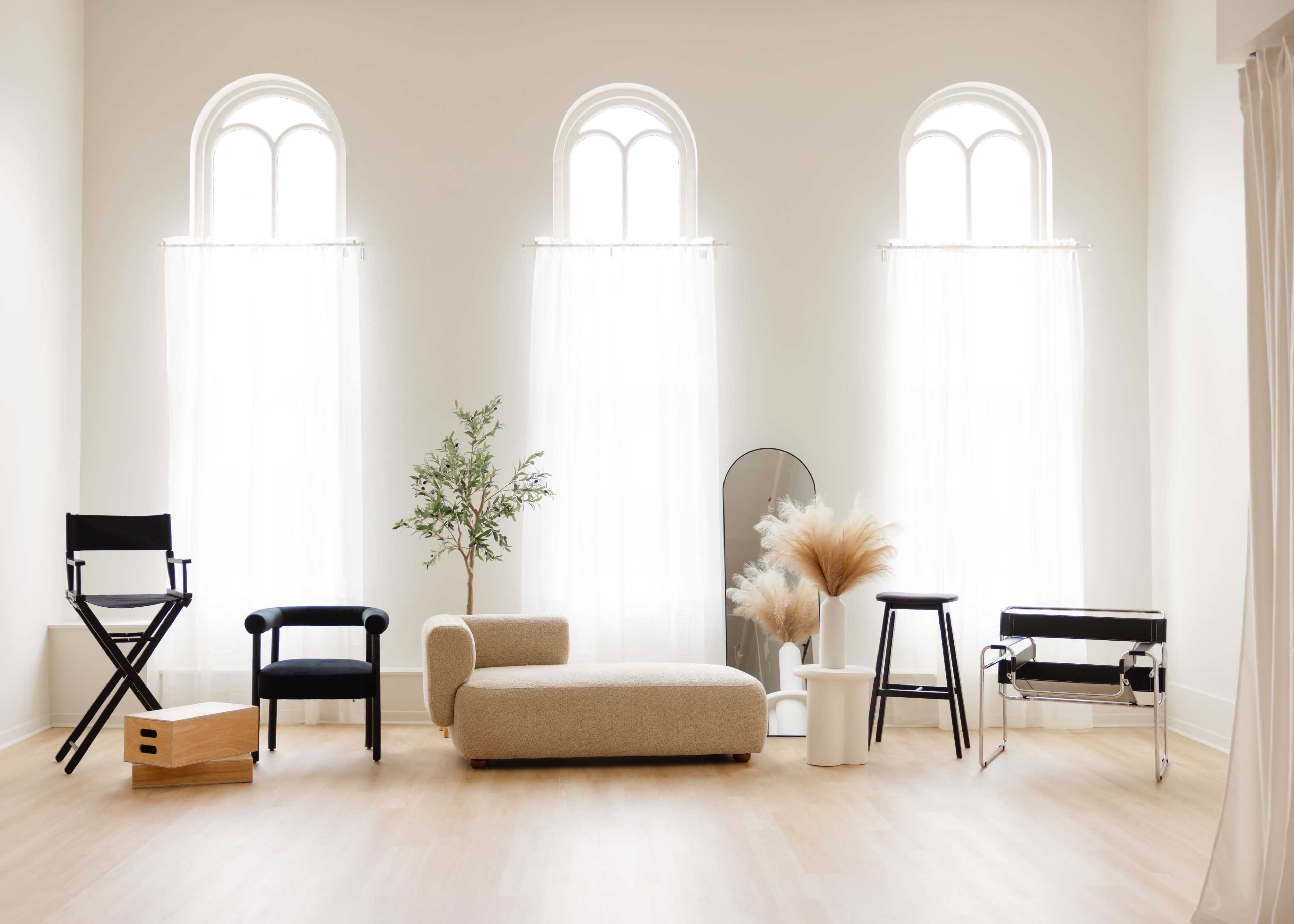 The image shows a modern interior space featuring various chairs, a potted plant, a mirror, and decorative greenery near large, arched windows.