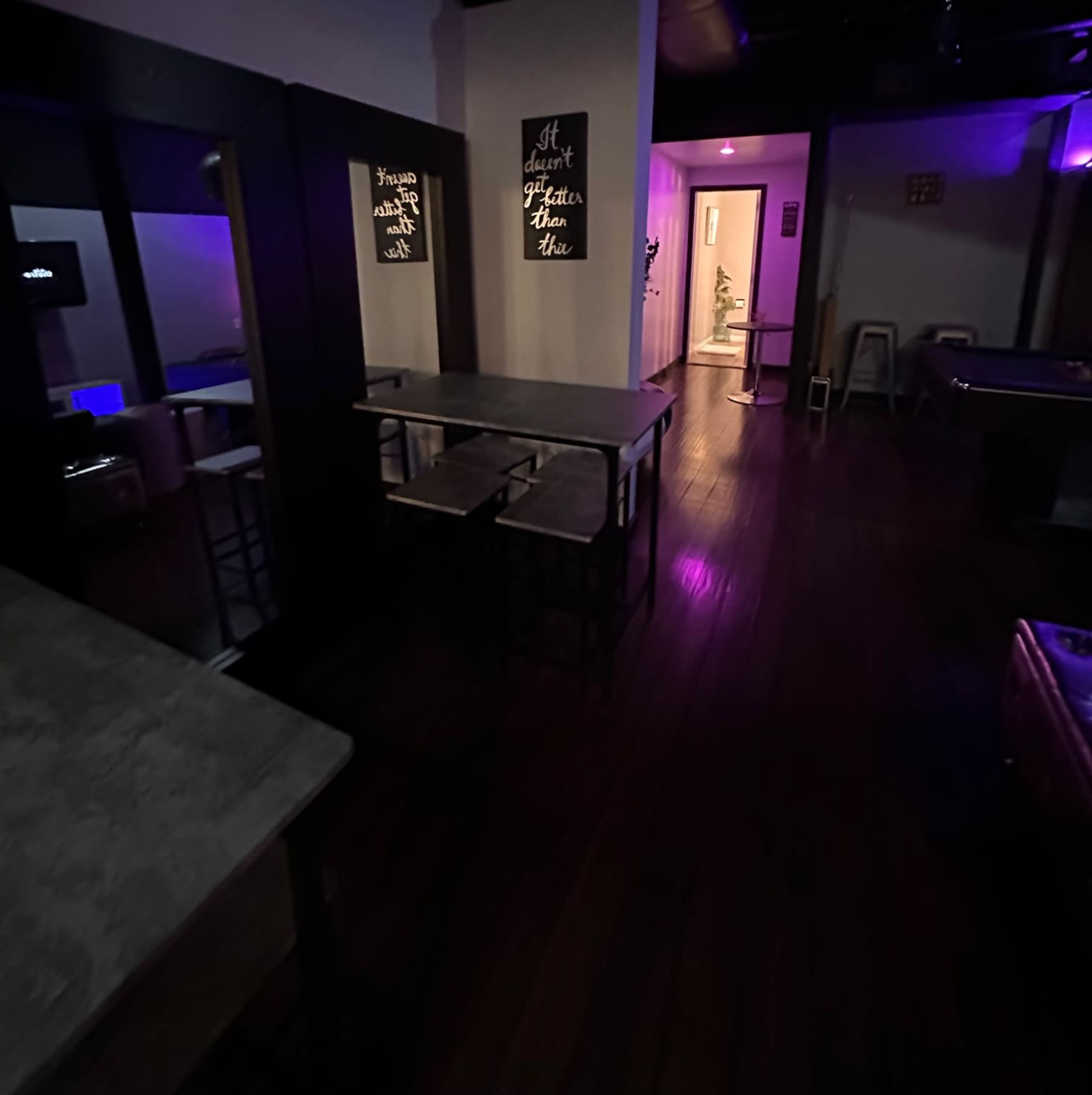 A dimly lit interior space with wooden flooring, featuring tables and two walls with illuminated text, leading to a brightly lit hallway.