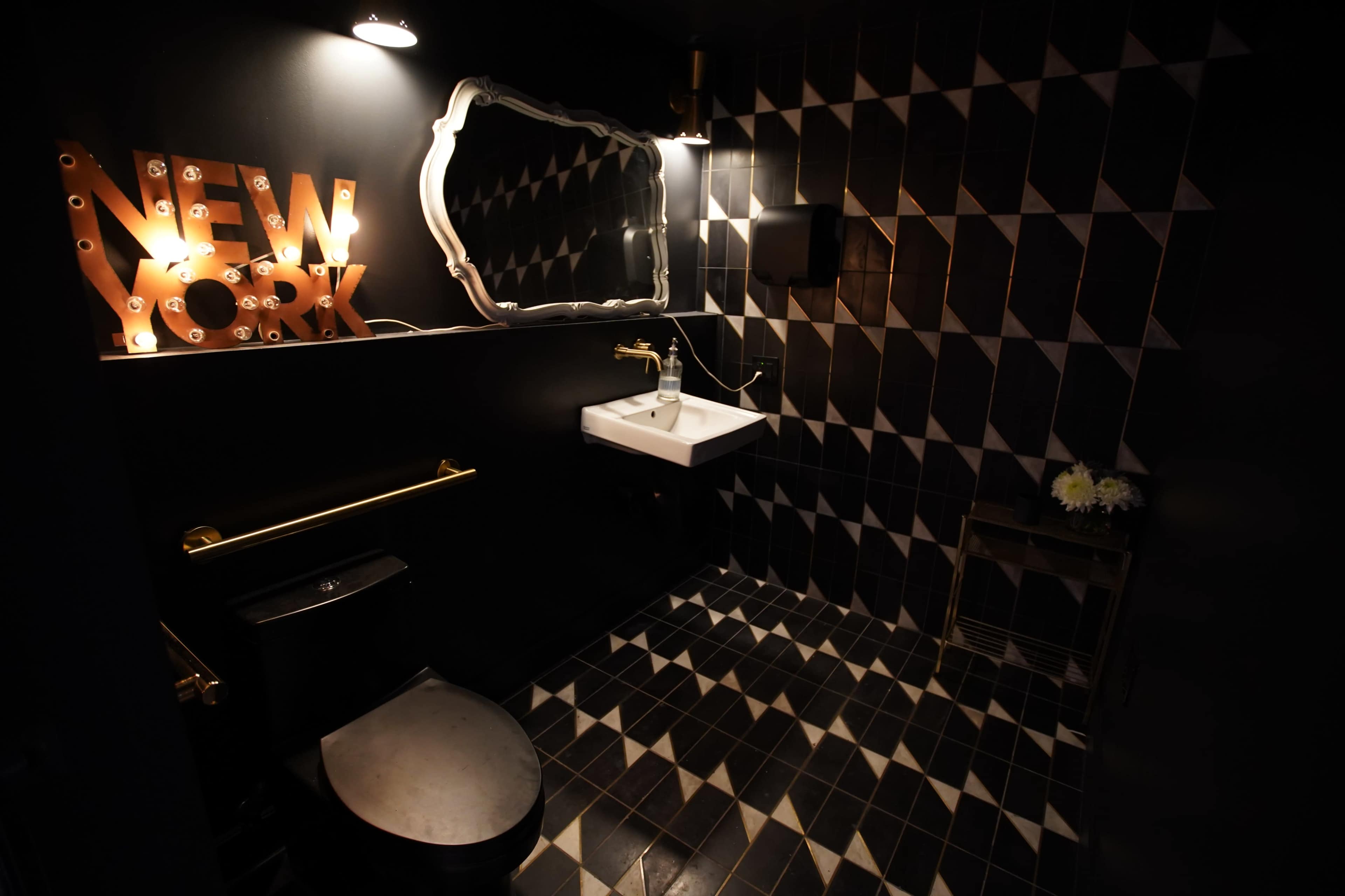 The image shows a modern bathroom featuring black and white diamond-patterned tiles, a white sink, a decorative mirror, and a lighted sign that reads "NEW YORK."
