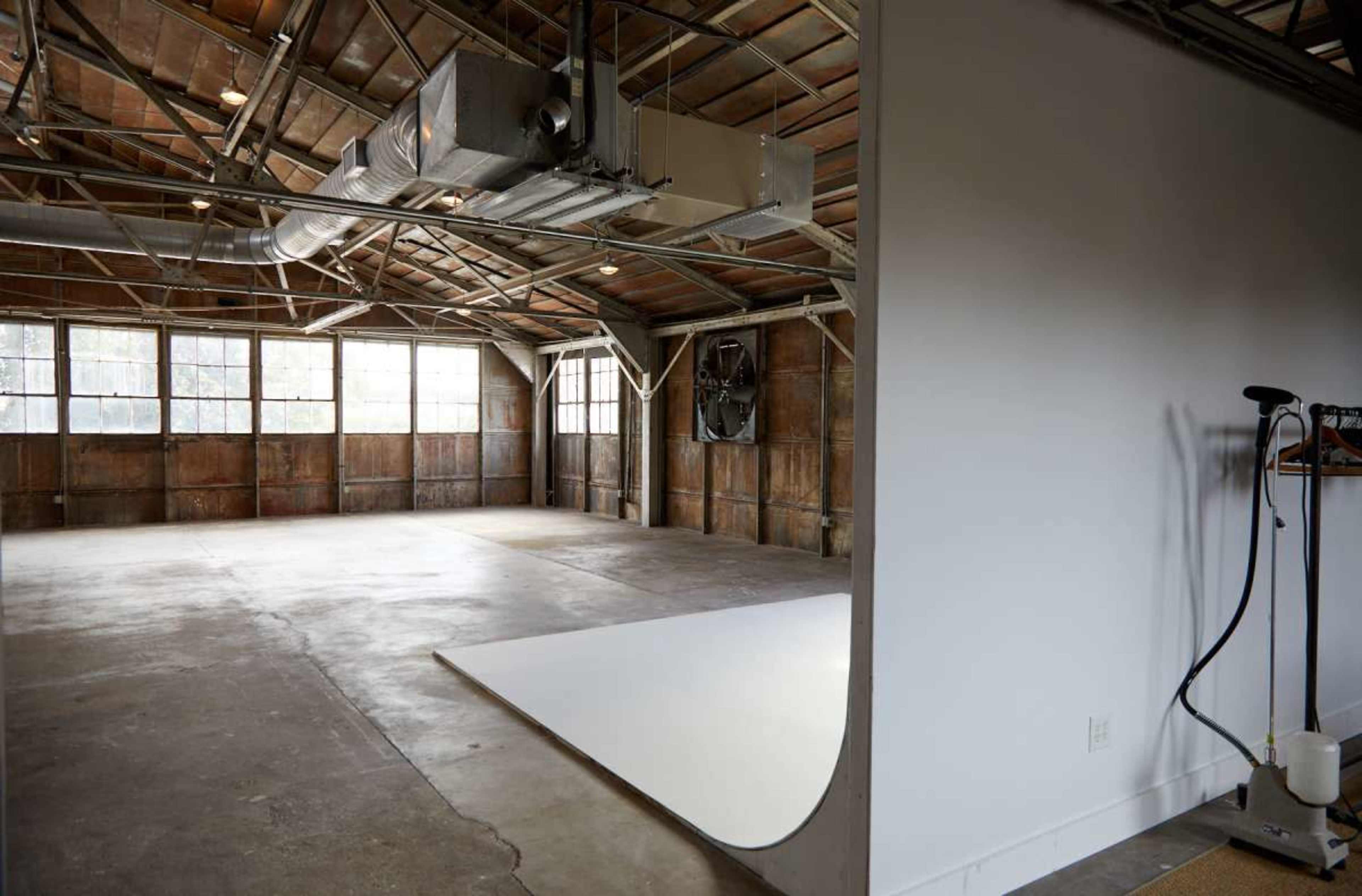 The image shows an empty, industrial-style room with exposed beams and large windows, featuring a partitioned area with a white backdrop and a steam cleaner in the corner.