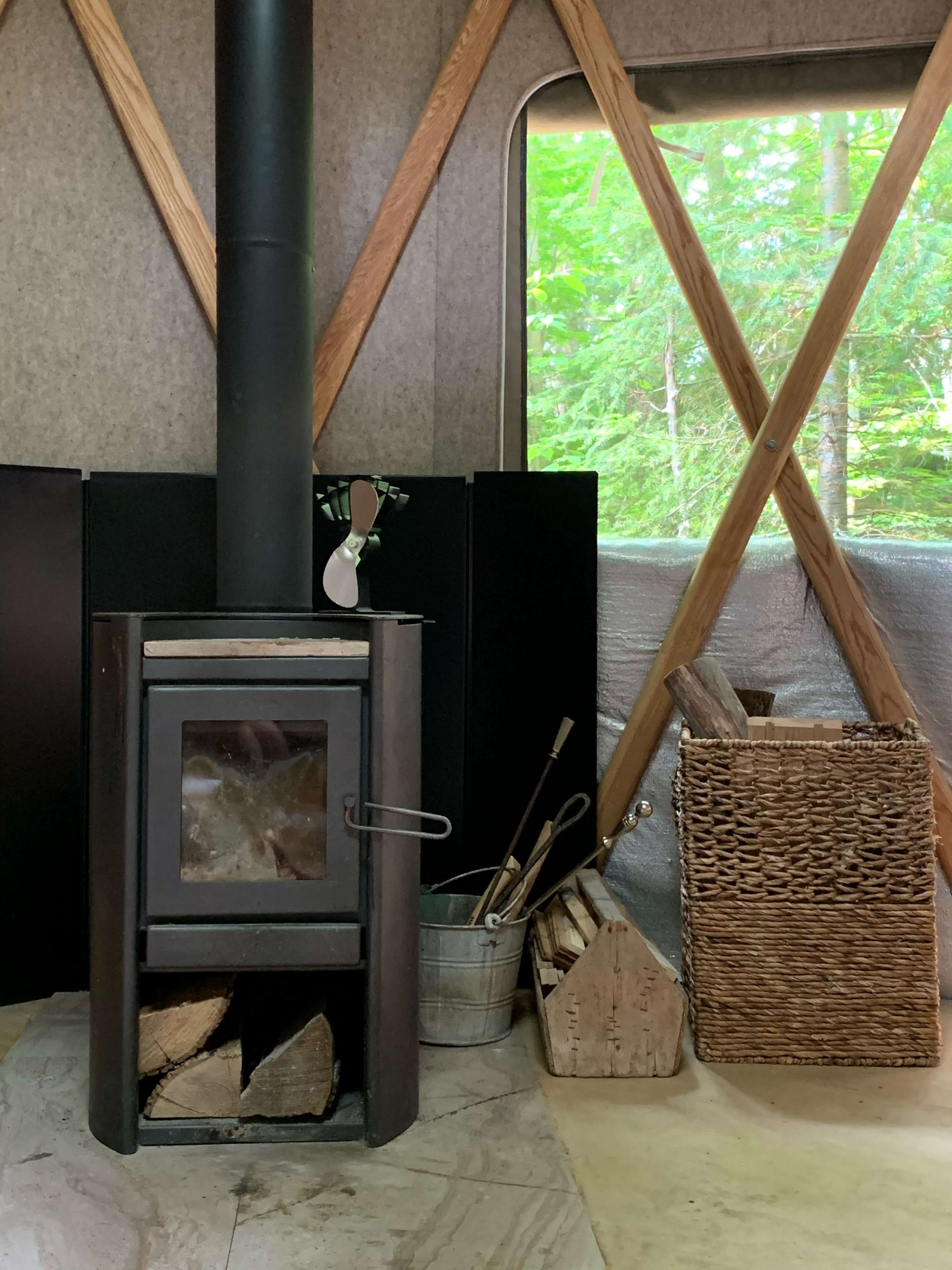 A wood stove beside a wicker basket filled with firewood in a yurt surrounded by greenery.
