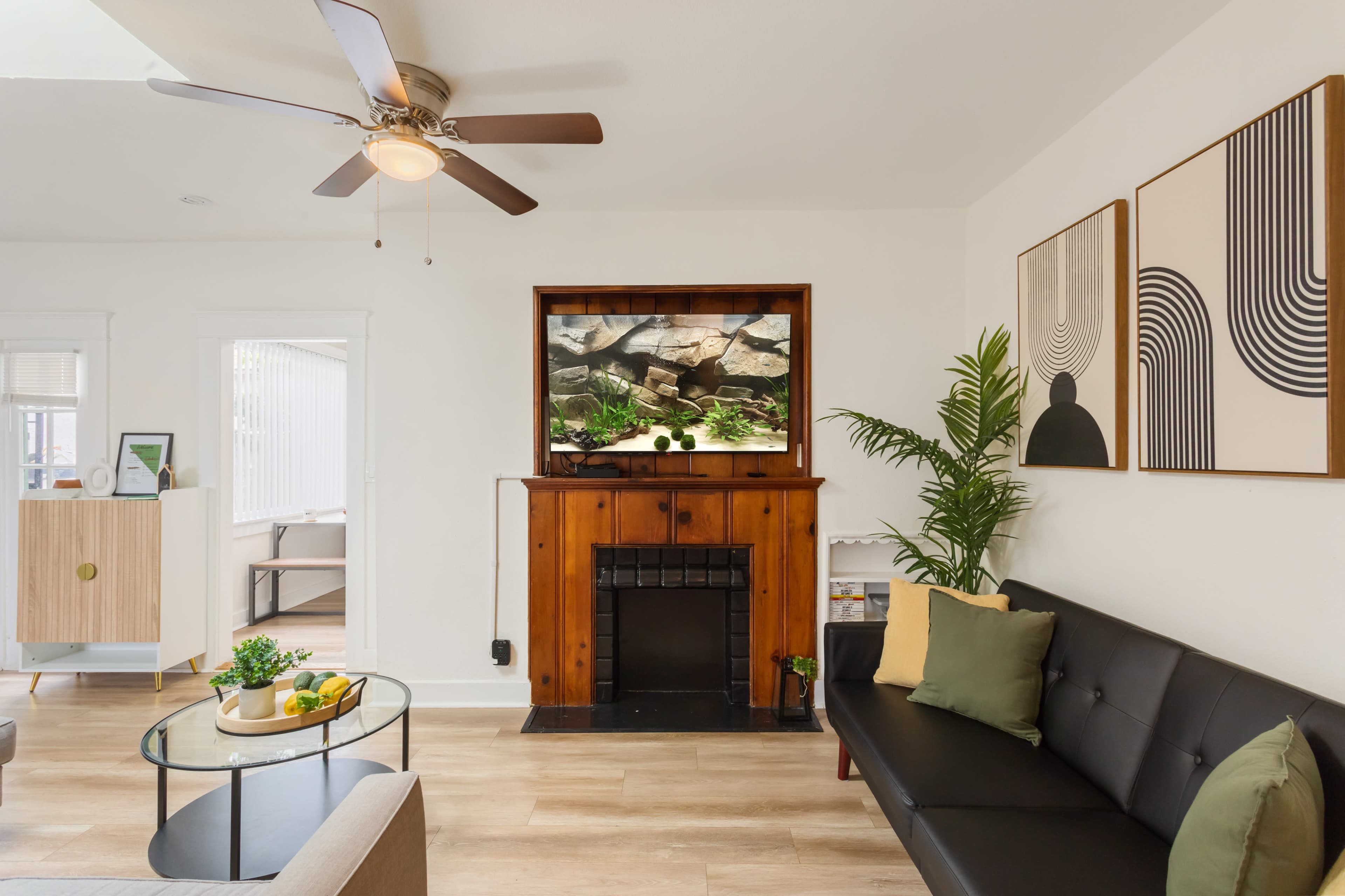 A modern living room features a black sofa, a glass coffee table with fruit, and a wall-mounted television above a wooden mantel.