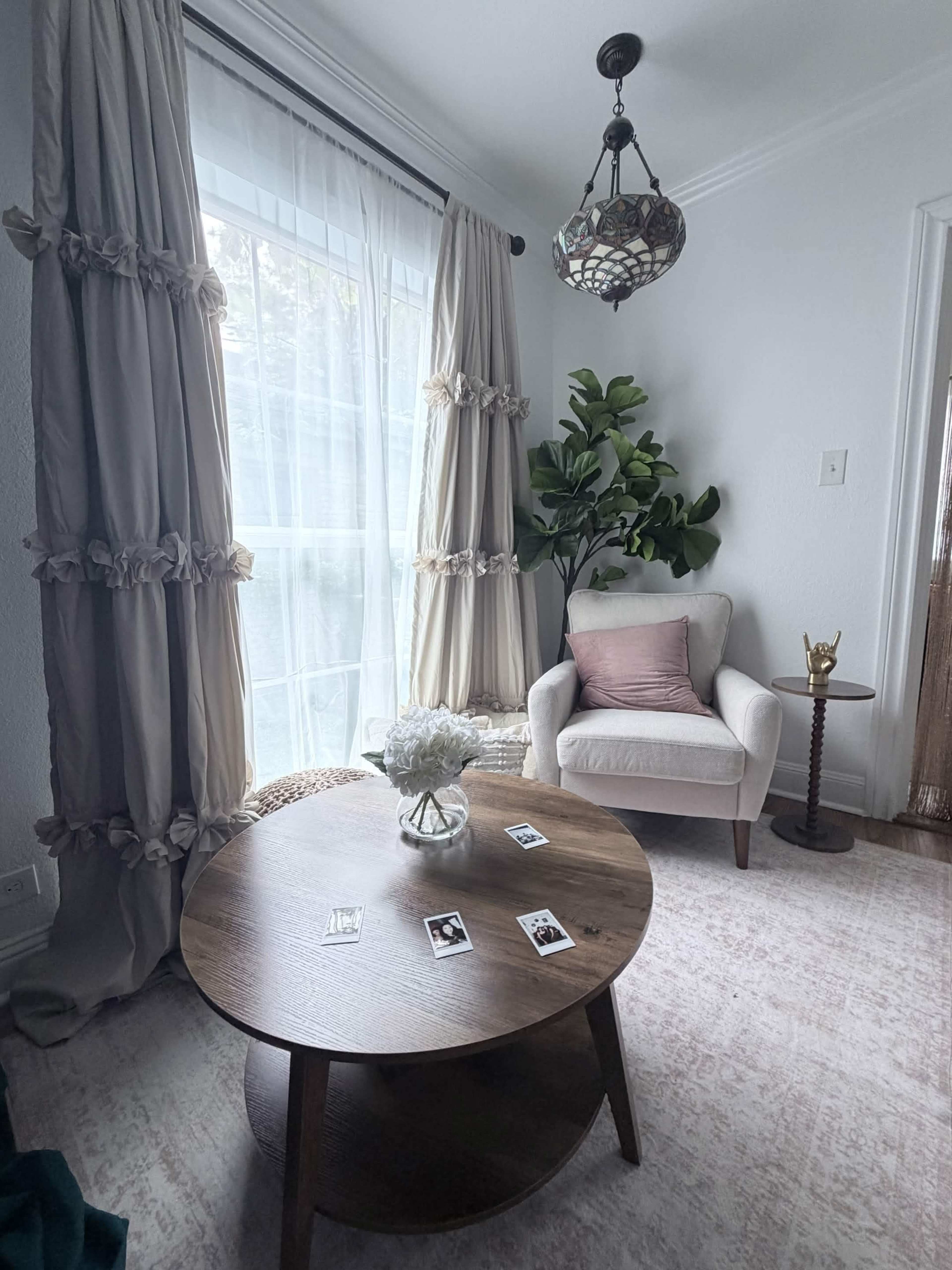 The image depicts a cozy sitting area featuring a light-colored armchair, a round wooden table with scattered photos, and floor-length curtains beside a large window adorned with a potted plant.