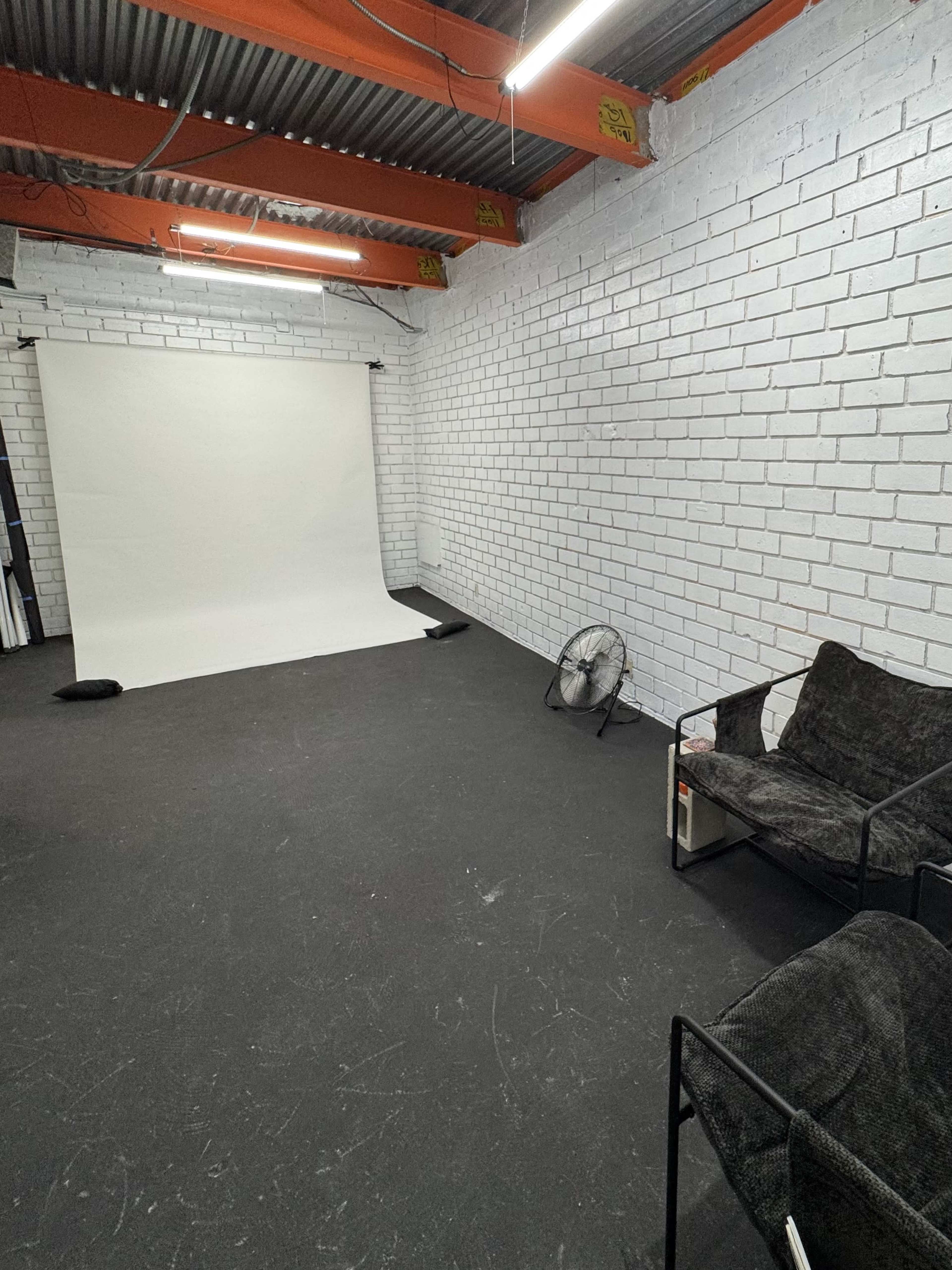 The image shows a minimalist studio space with a white backdrop, a fan, and a black sofa against the brick walls.