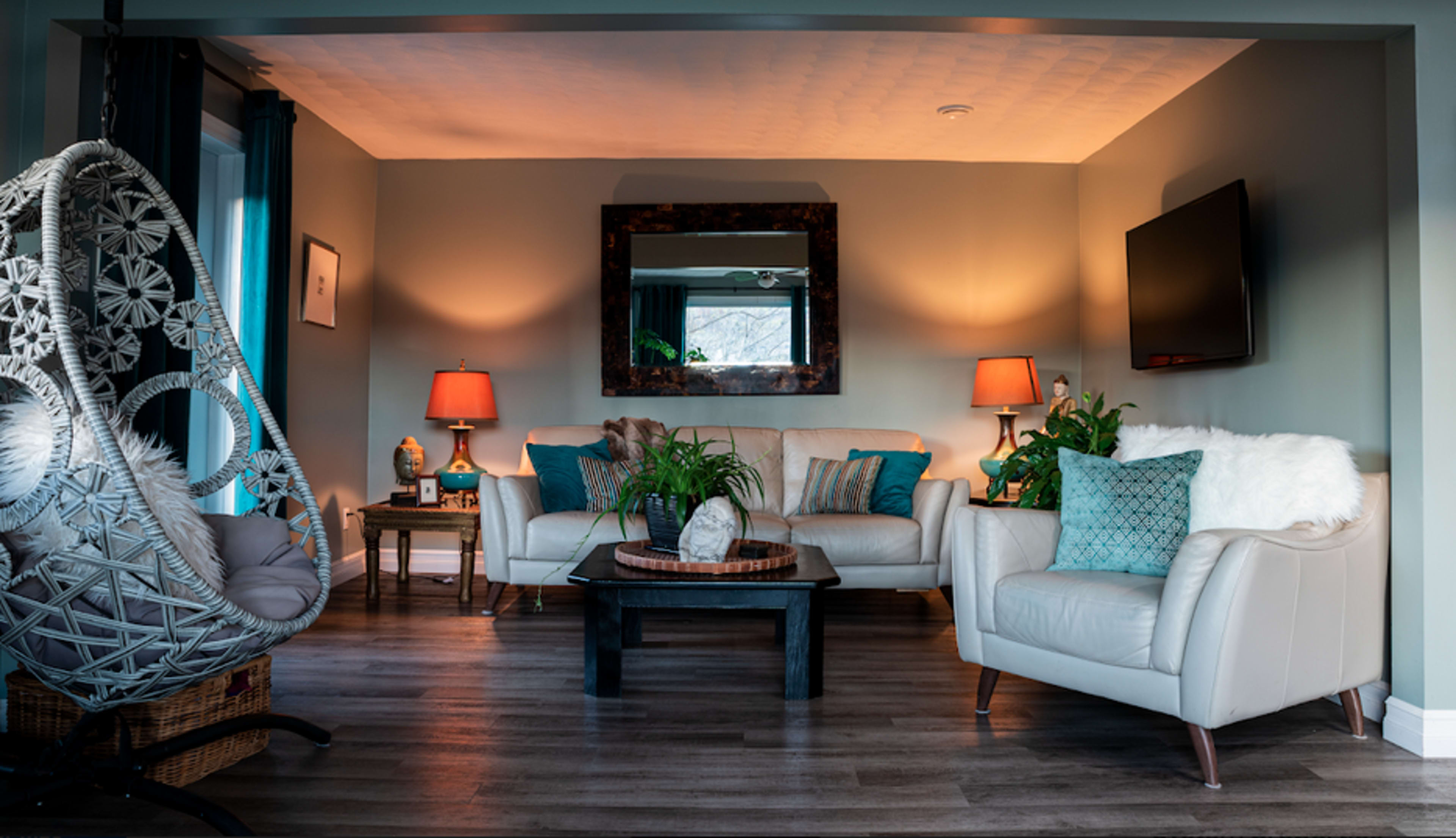 A cozy living room features a white leather sofa, an armchair, a swinging chair, and plants, illuminated by table lamps and a large mirror.