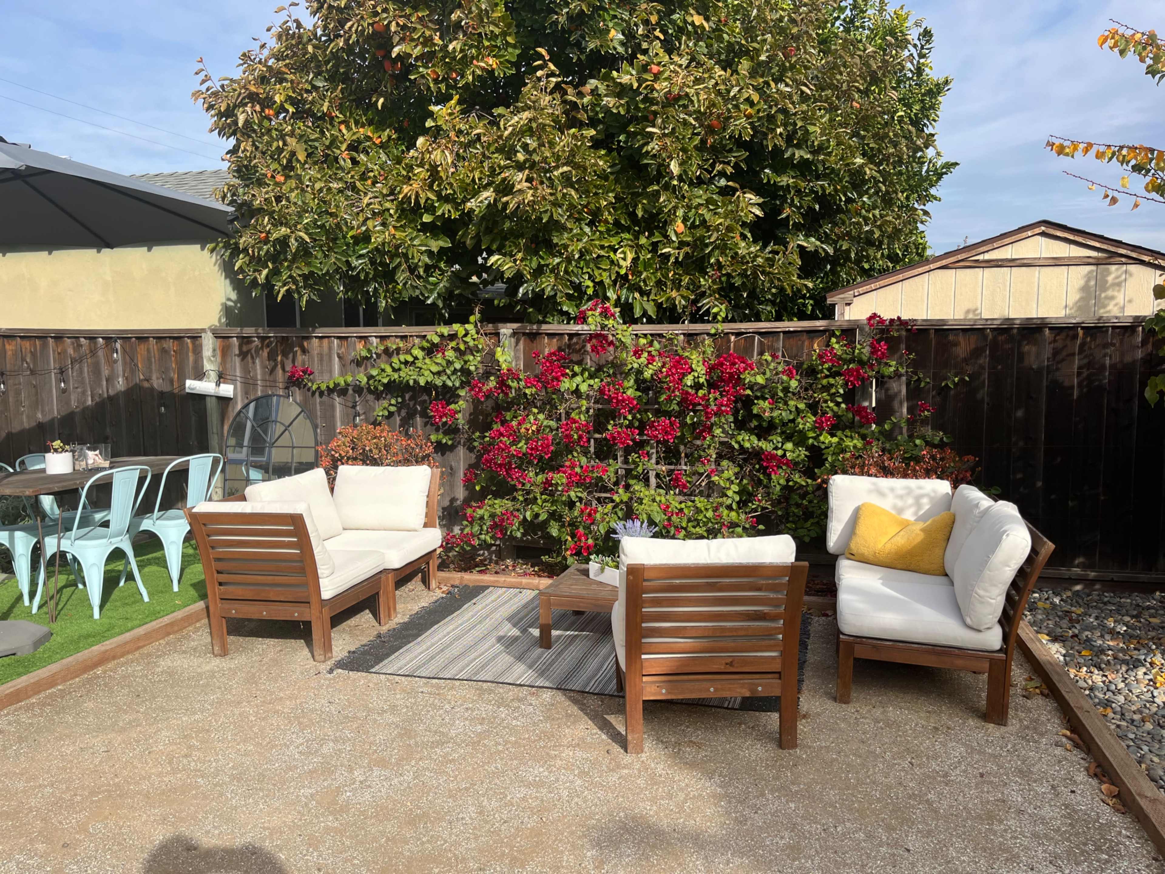 The image shows a small outdoor seating area with four wooden lounge chairs and a small table surrounded by flowering plants and a gravel surface.