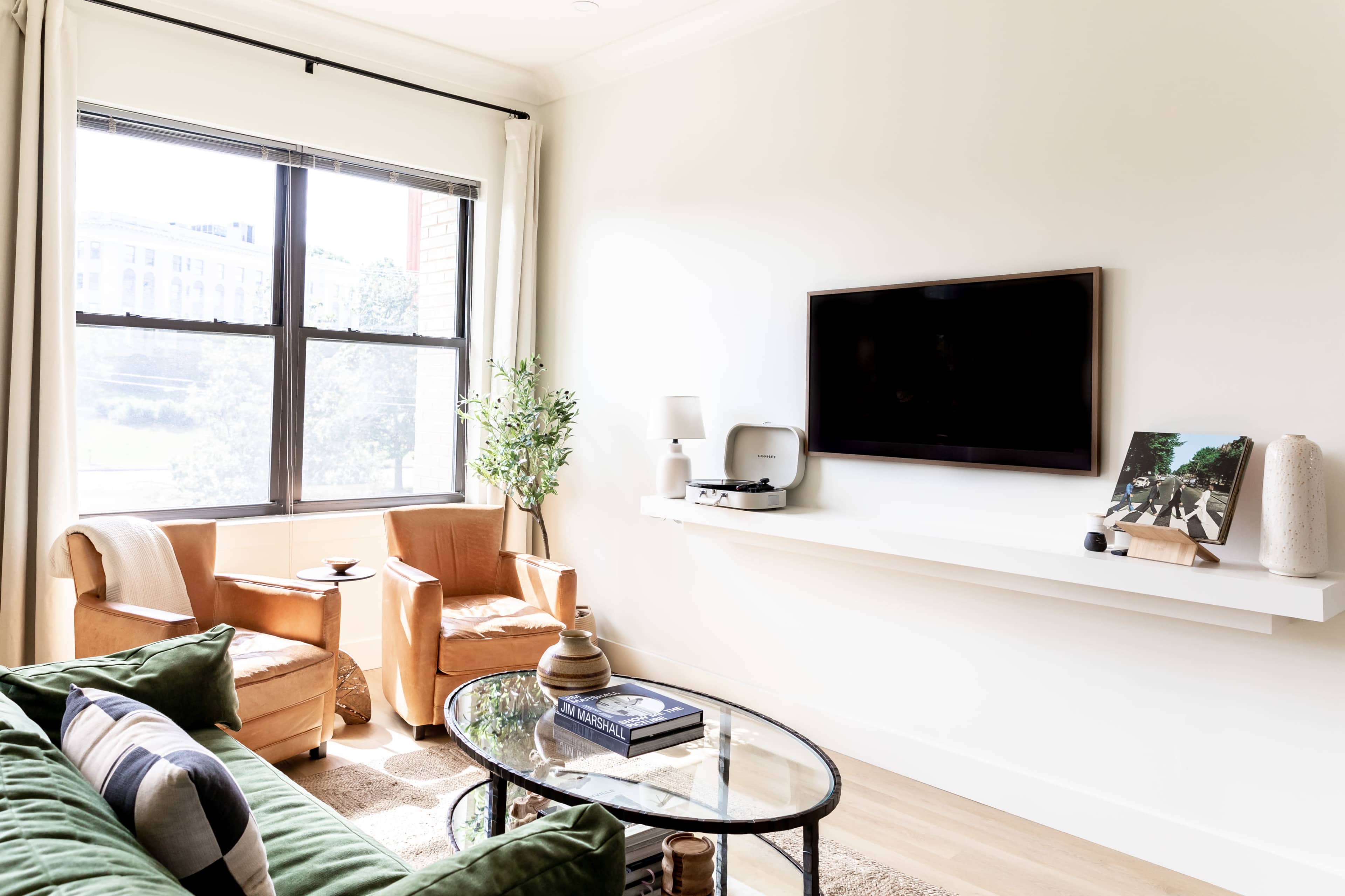 A modern living room features a green sofa, two brown chairs, a coffee table, and a mounted television on a white wall.