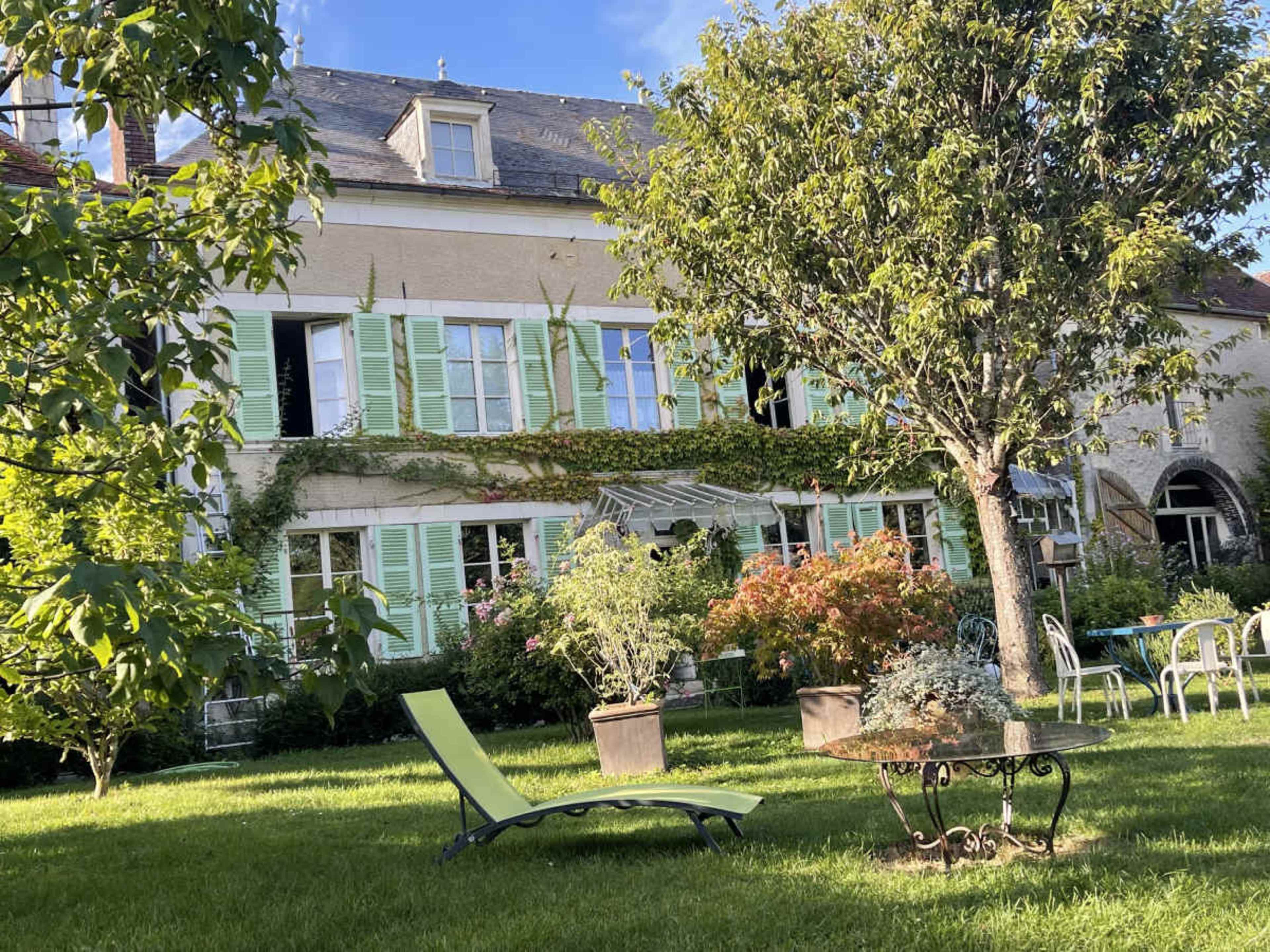The image shows a garden area with a green lounge chair and various plants in front of a house featuring light blue shutters and a well-maintained lawn.