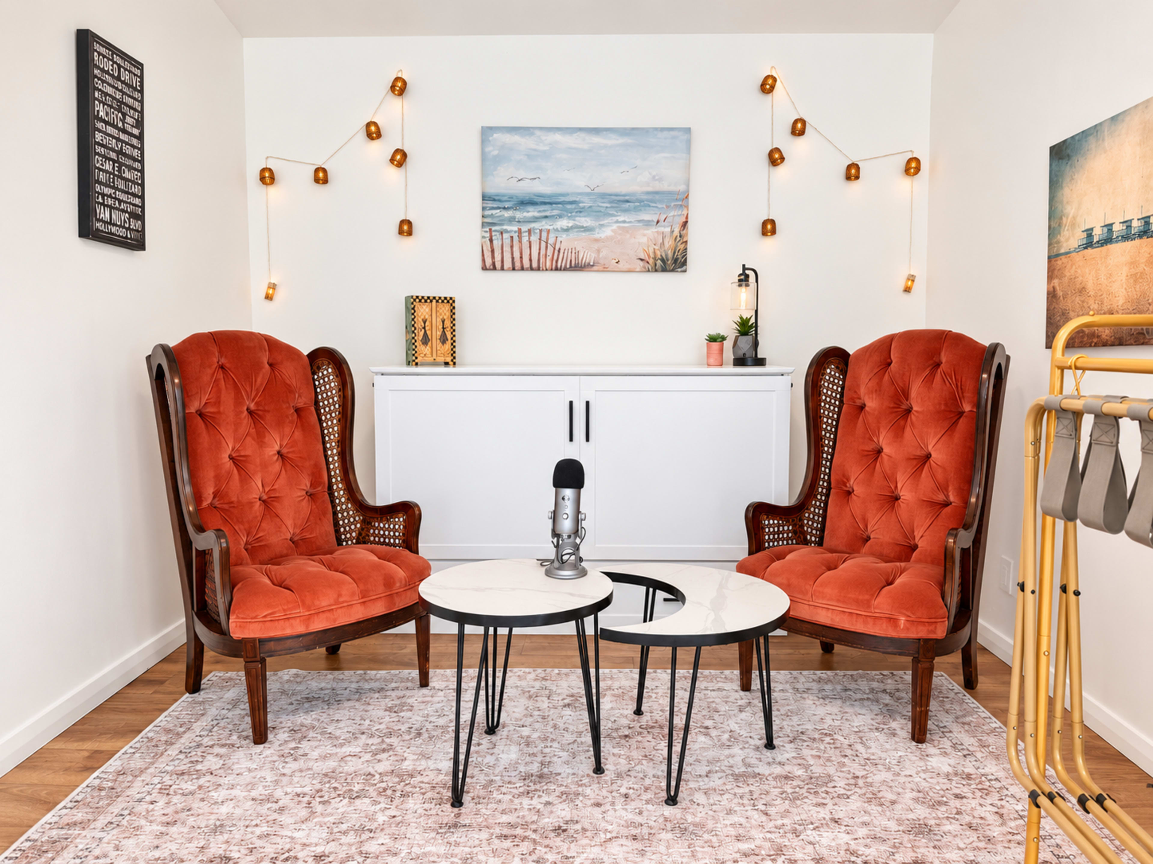 The image features a cozy room with two orange upholstered armchairs facing a marble-topped coffee table, adorned with string lights and a painting of a beach on the wall.