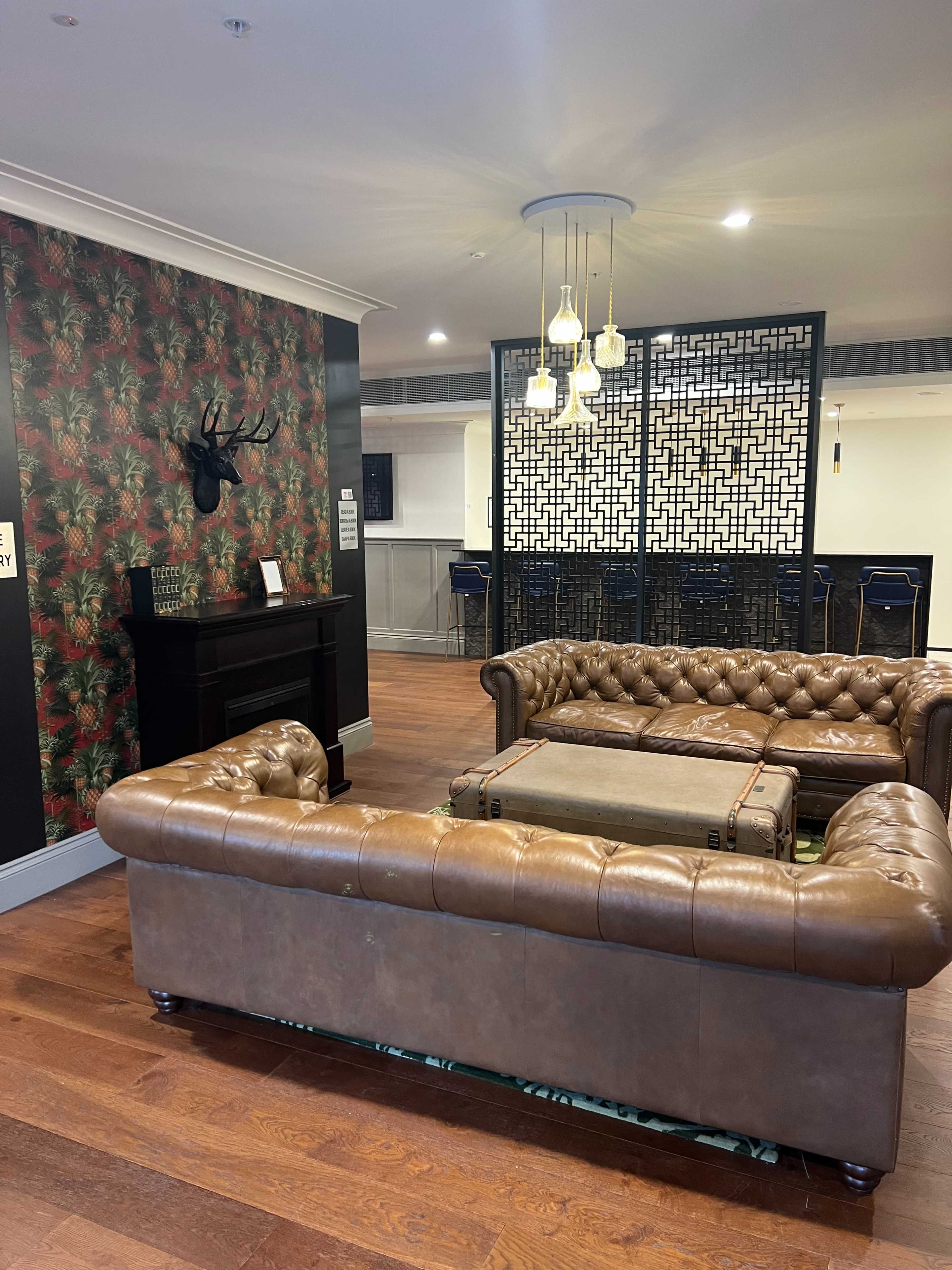 A lounge area featuring two brown leather tufted sofas, a patterned wallpaper with foliage, and hanging light fixtures.