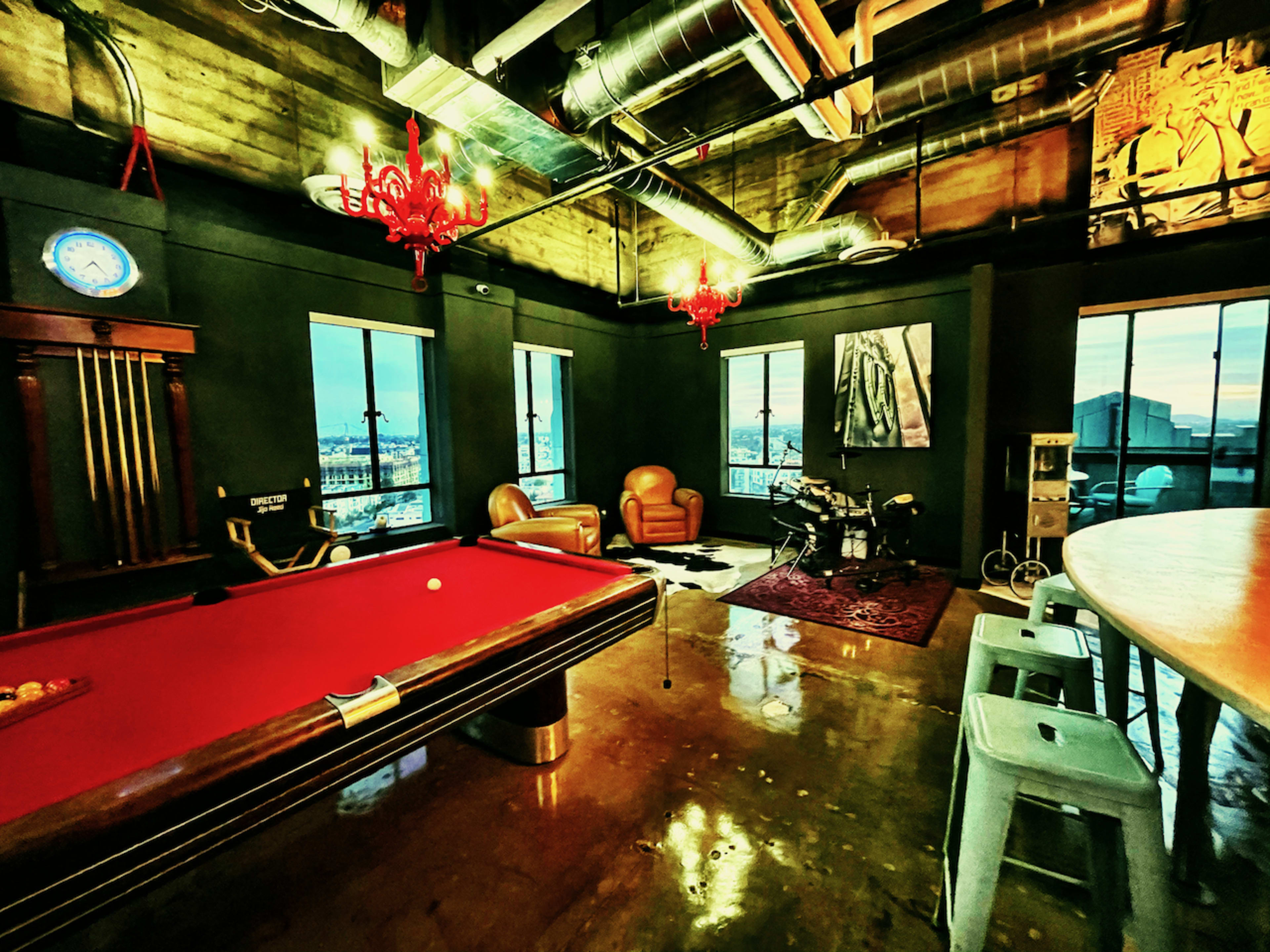 The image shows a modern game room featuring a pool table, leather chairs, a drum set, and large windows with an urban view.