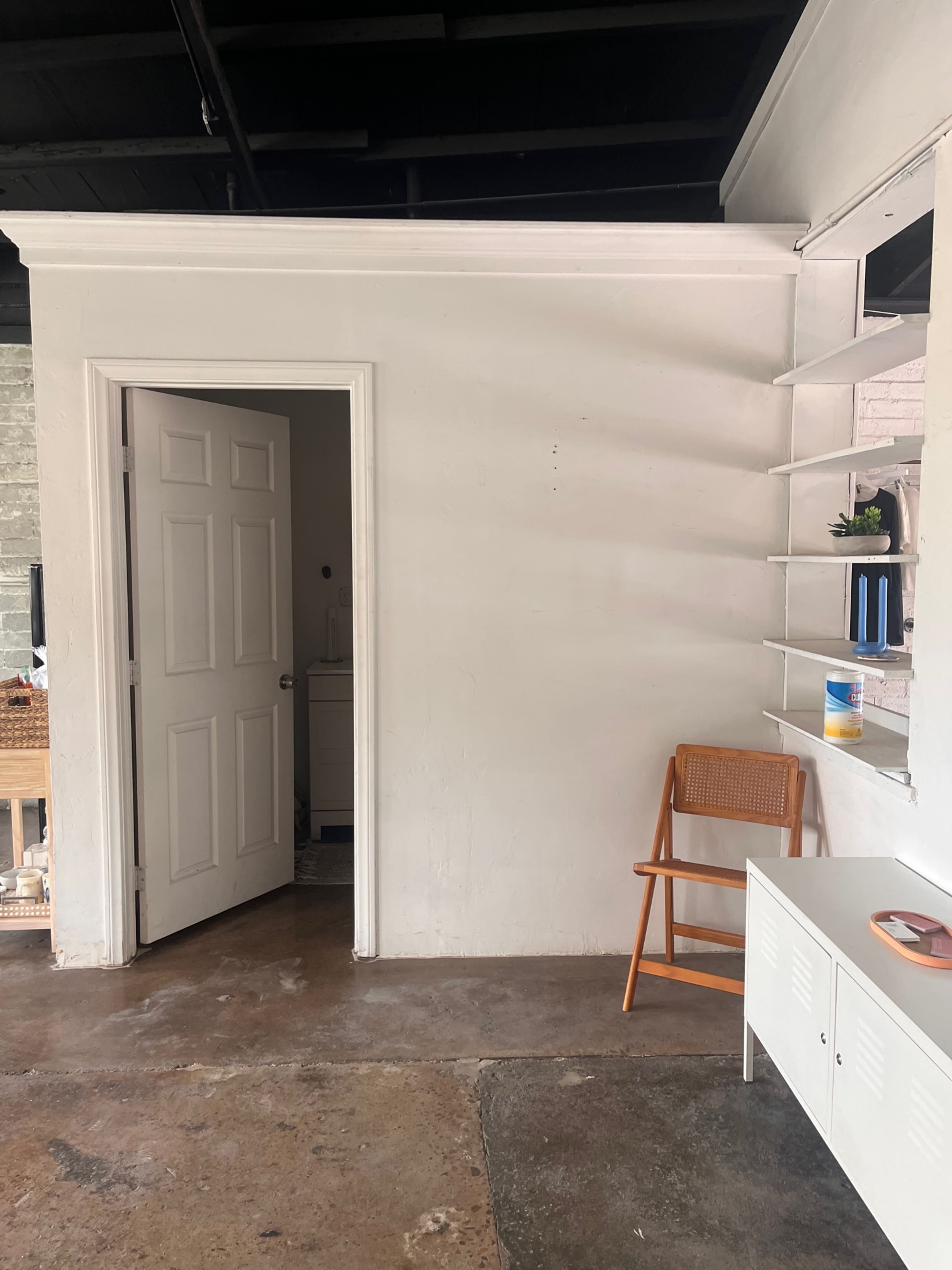 A small room features a white wall with a partially open door, a wooden chair, and a shelf with decorative items.