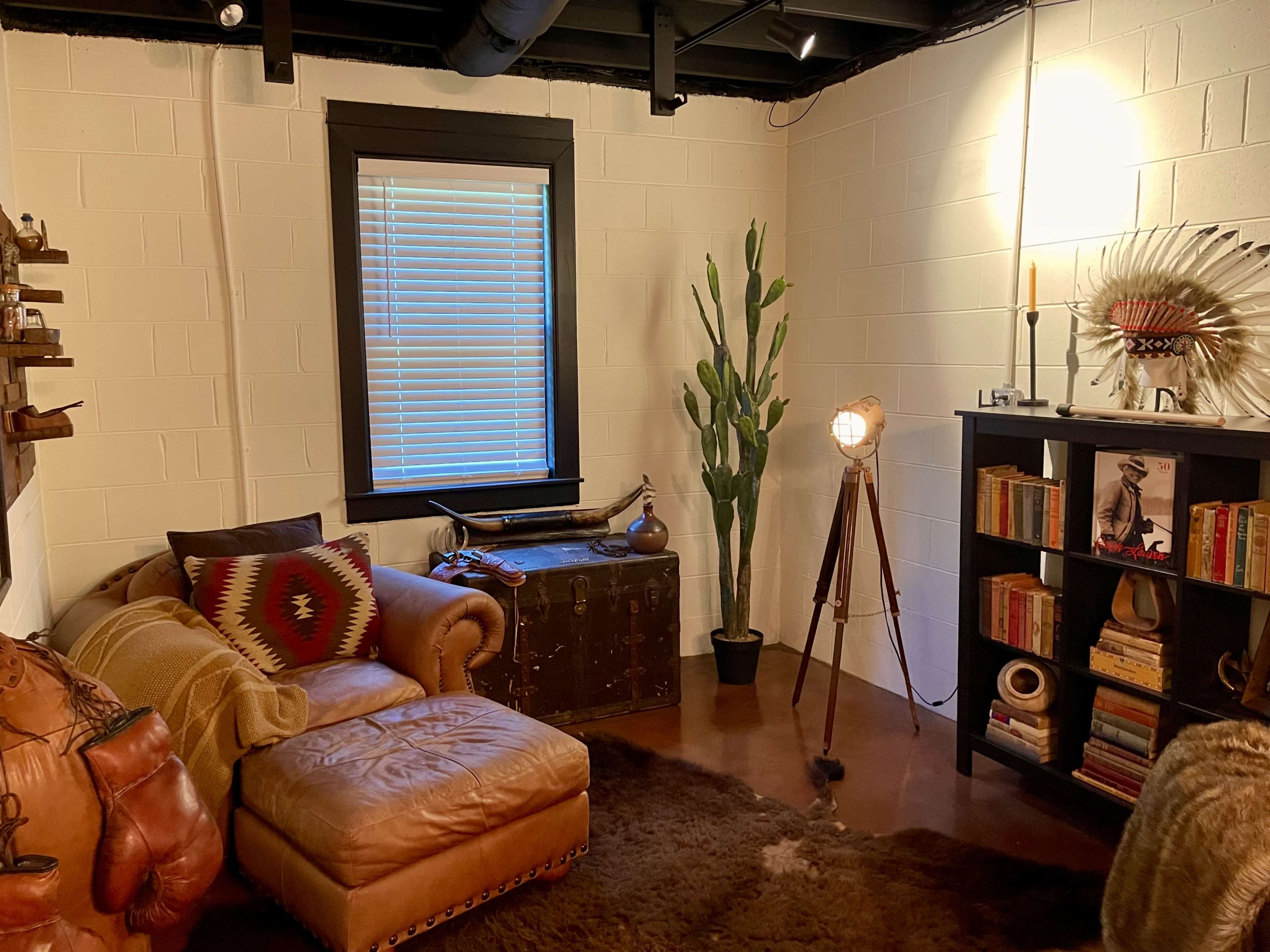 A cozy room featuring a leather chaise lounge, a large plant, a bookshelf filled with books, and a vintage-style lamp.