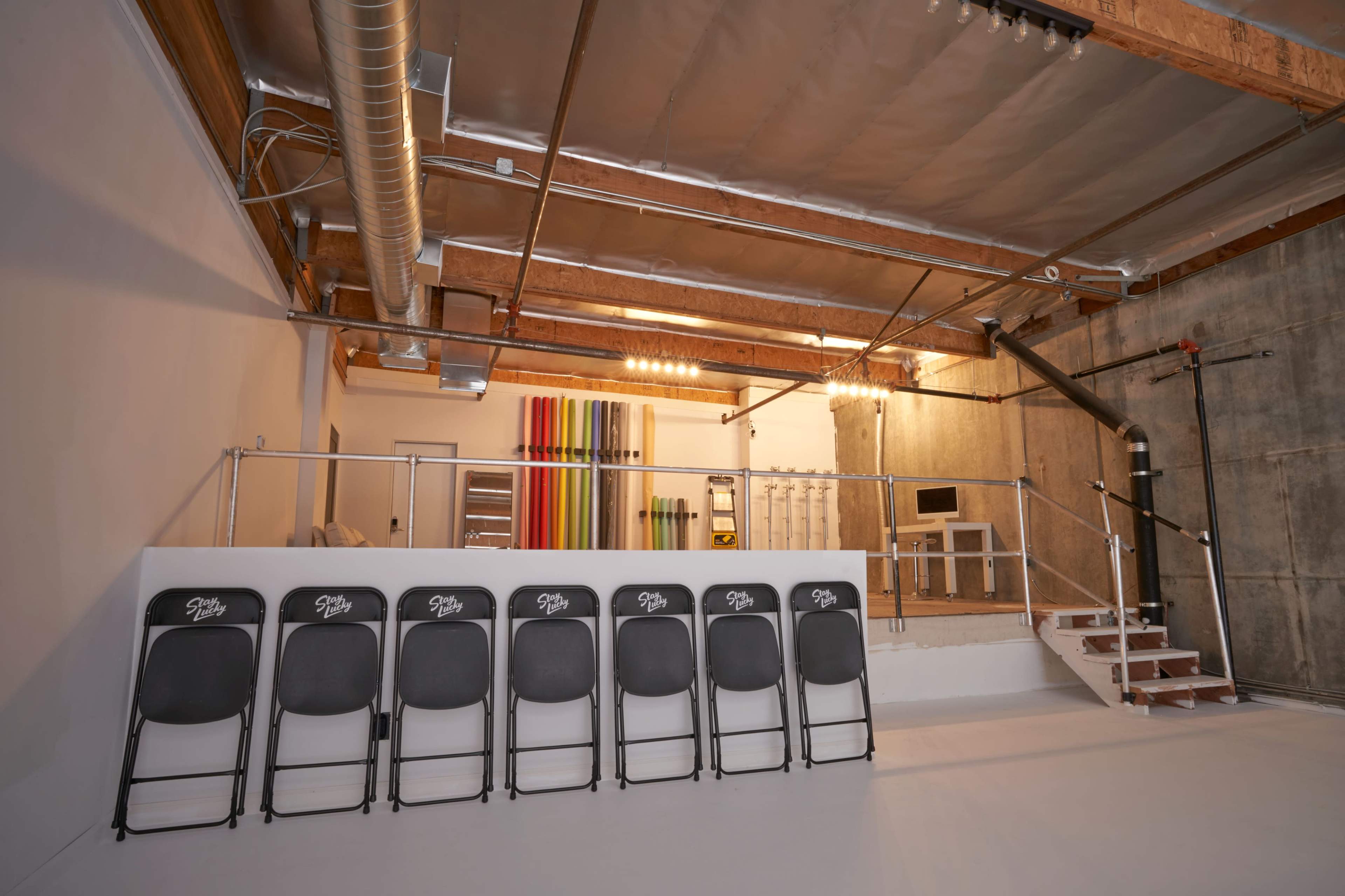 A minimalist interior space with a row of black folding chairs facing a staircase and a section featuring colorful rolled materials on the wall.