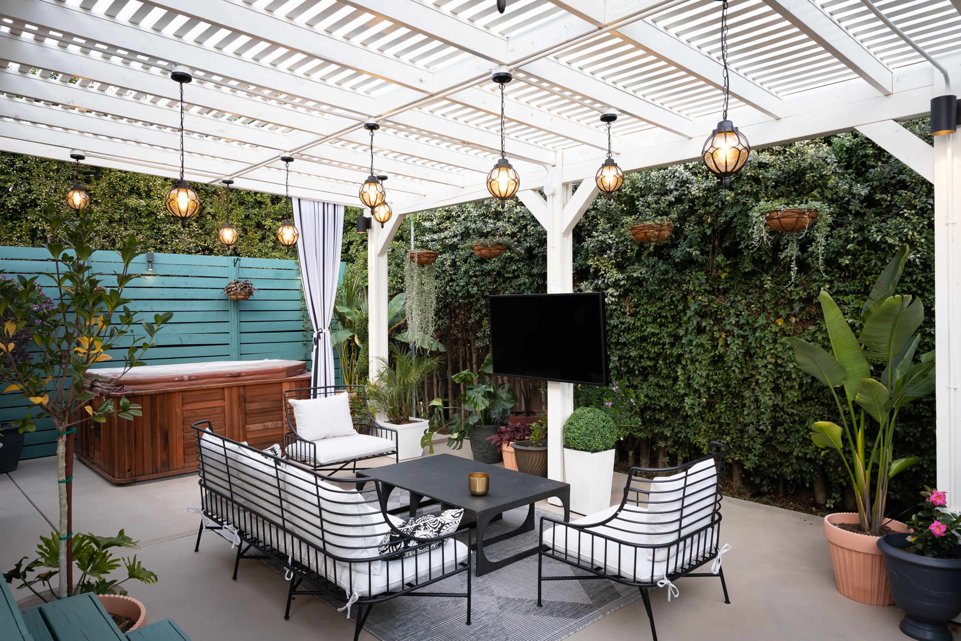A shaded outdoor patio features a seating area with a black table and white chairs, a hot tub in the background, and string lights overhead.