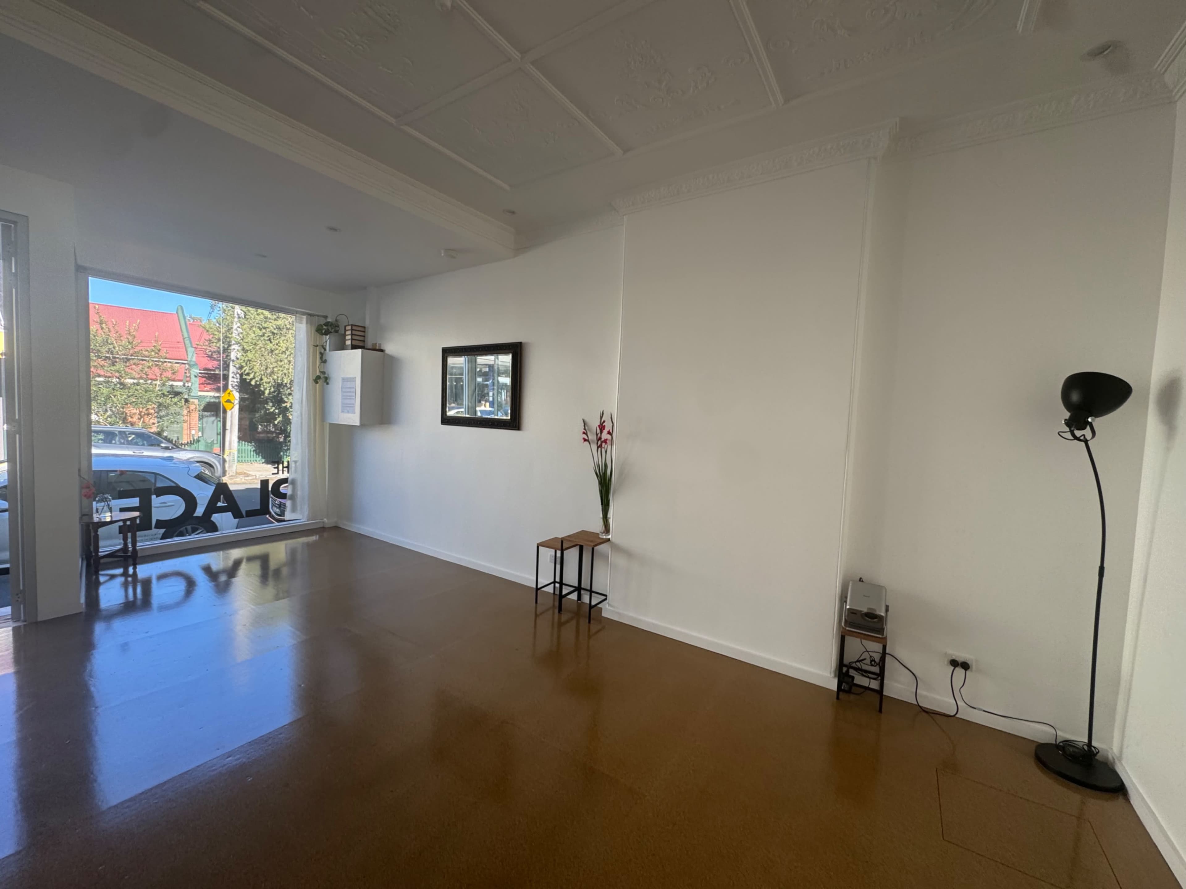 The image shows a spacious, empty interior with polished floors, a small table, a lamp, and a view of the street through a large window.