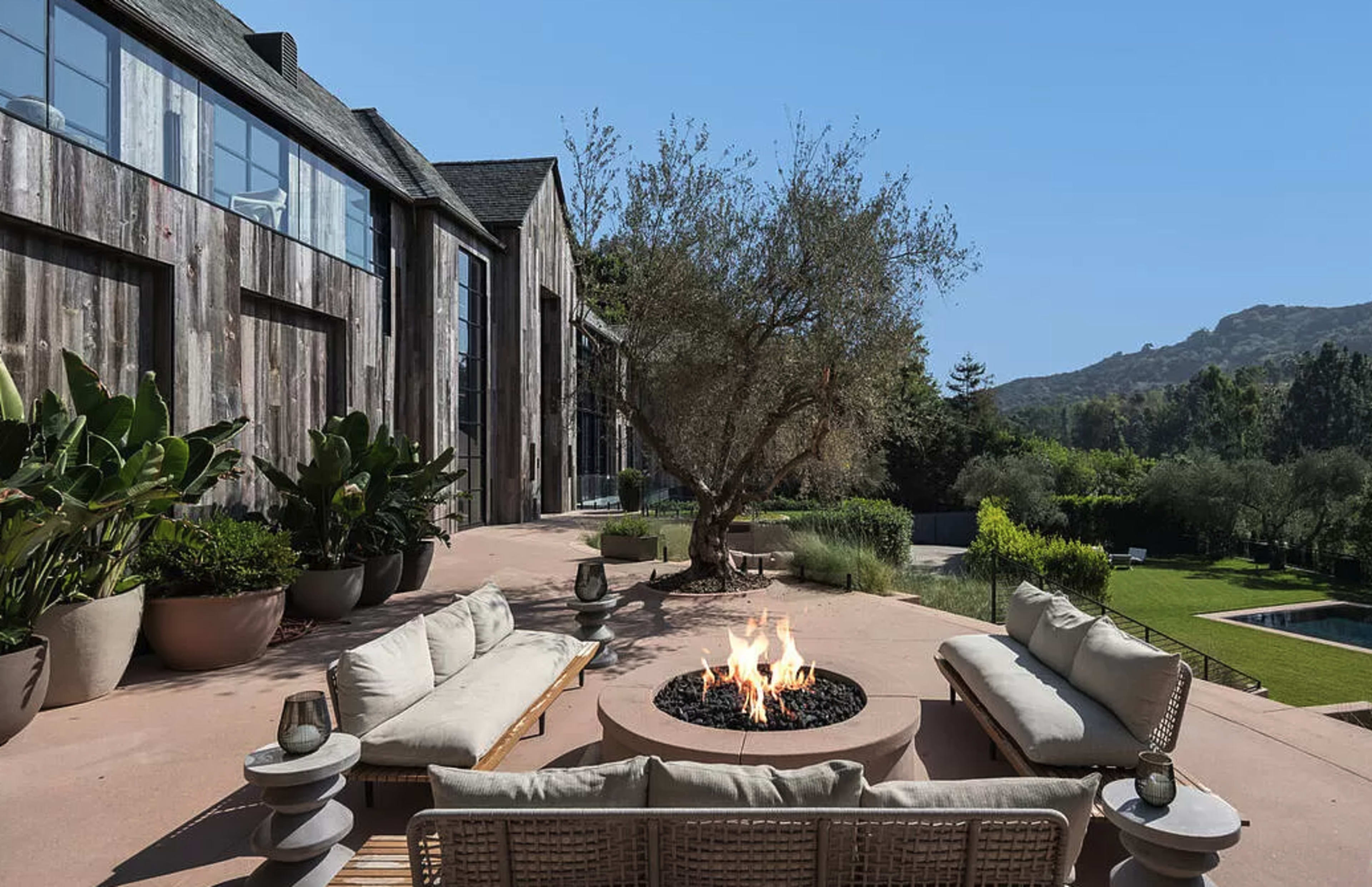 A circular fire pit with flames is surrounded by outdoor seating, set against a backdrop of a modern home and manicured greenery.