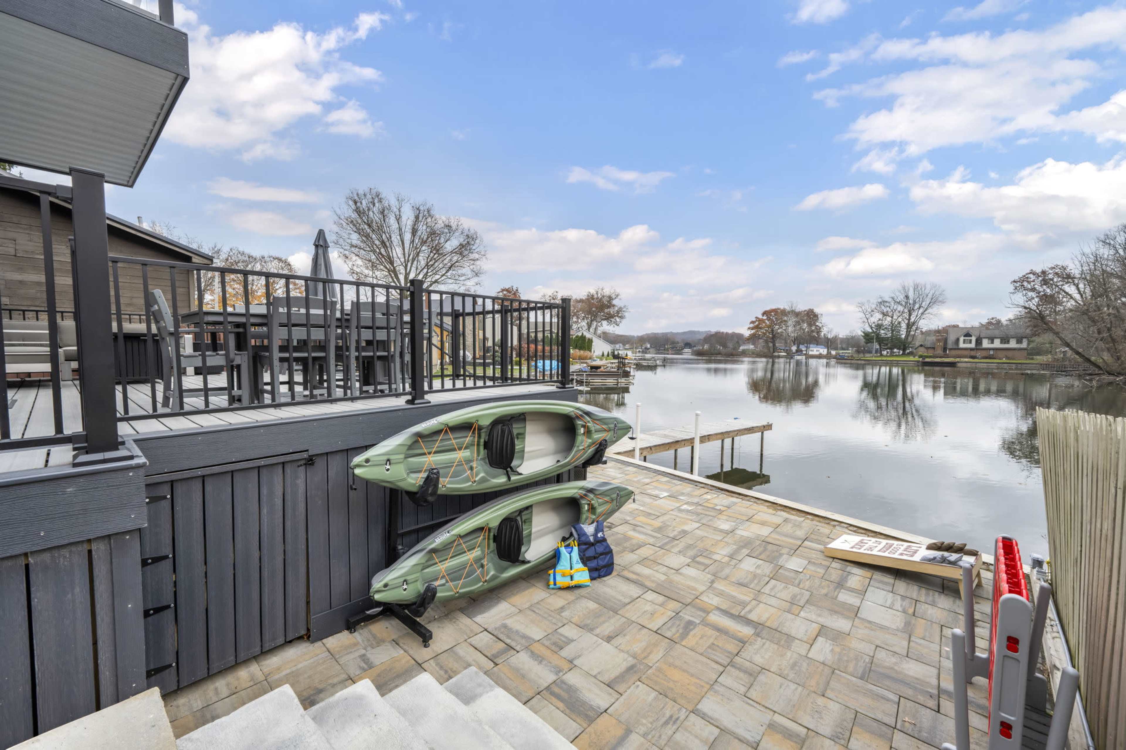 The image shows a patio area beside a calm river, featuring two kayaks stacked against the wall and a staircase leading down to the water.
