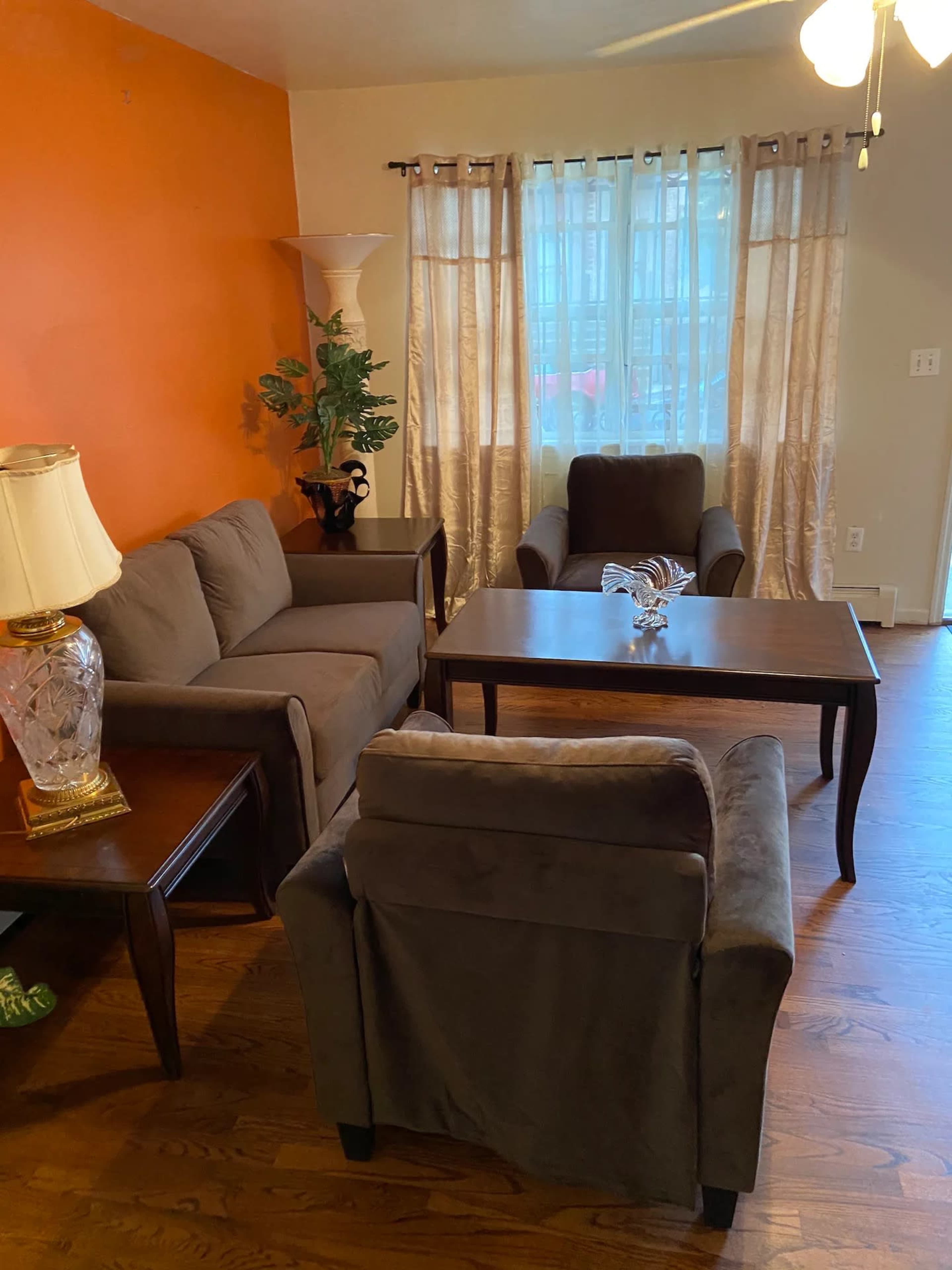A living room furnished with a brown sofa, a pair of armchairs, a coffee table, and a lamp, alongside a window with sheer curtains.
