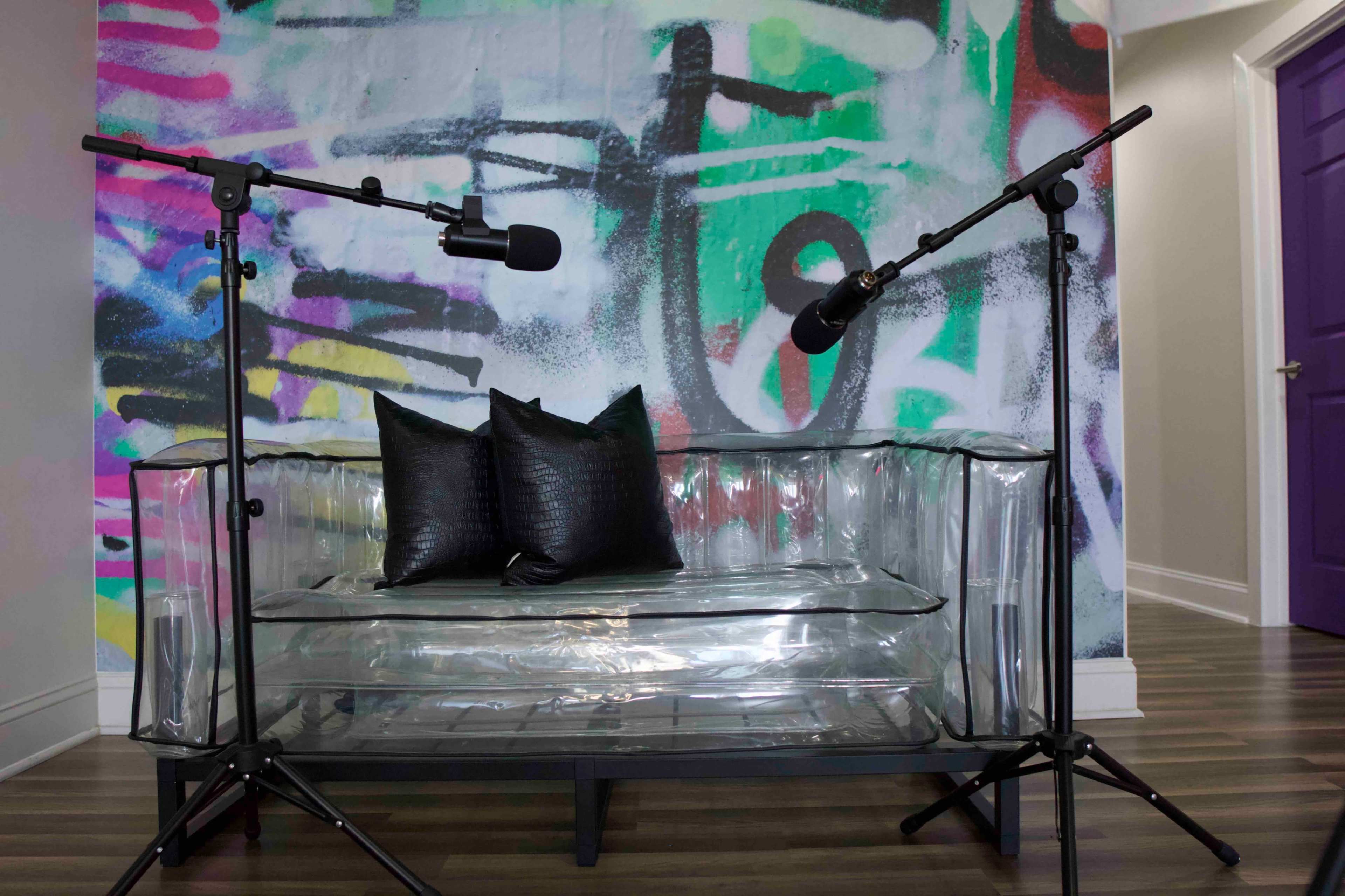 A transparent acrylic couch with two black pillows is positioned in front of a colorful graffiti-style wall, flanked by two microphone stands.