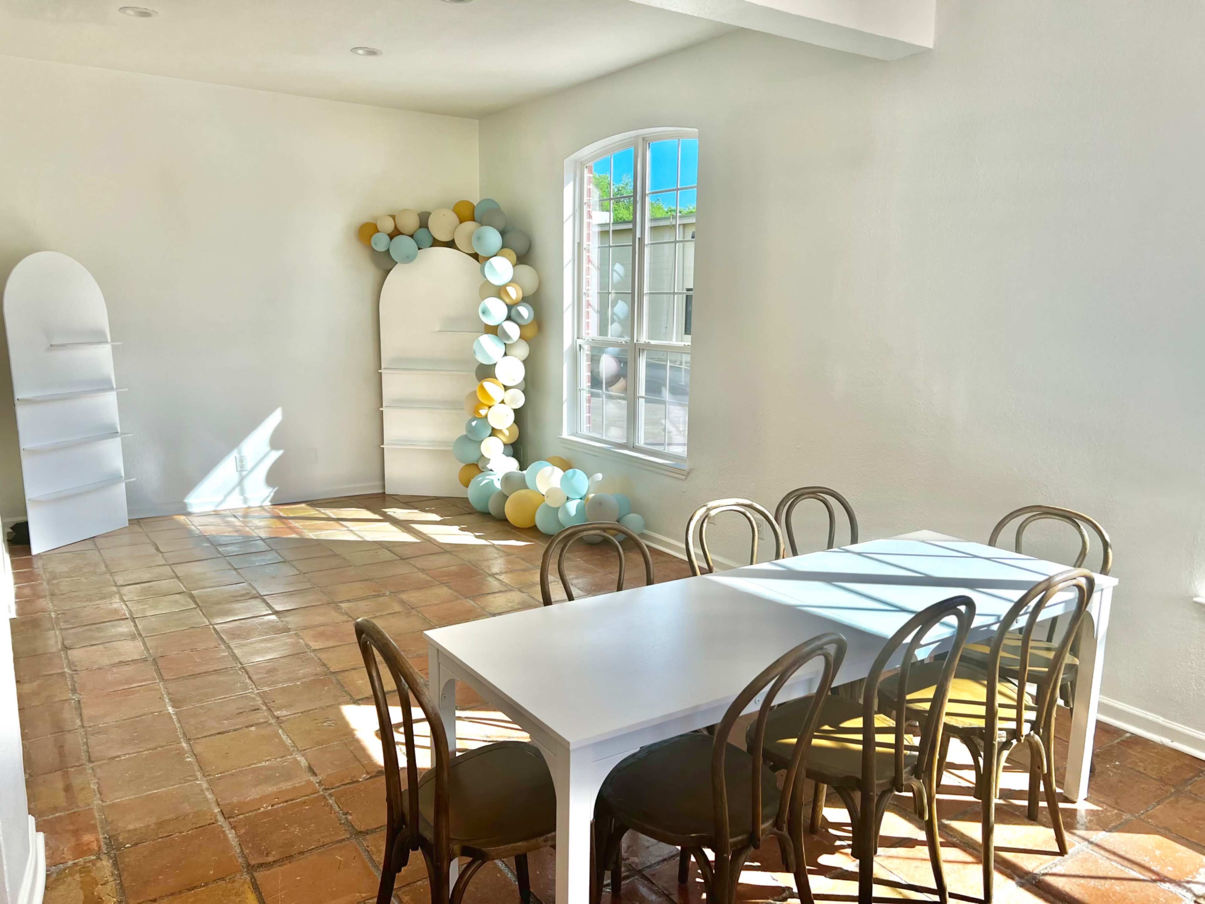 The image shows a well-lit room with a white table, six wooden chairs, and a decorative arrangement of balloons against the wall.
