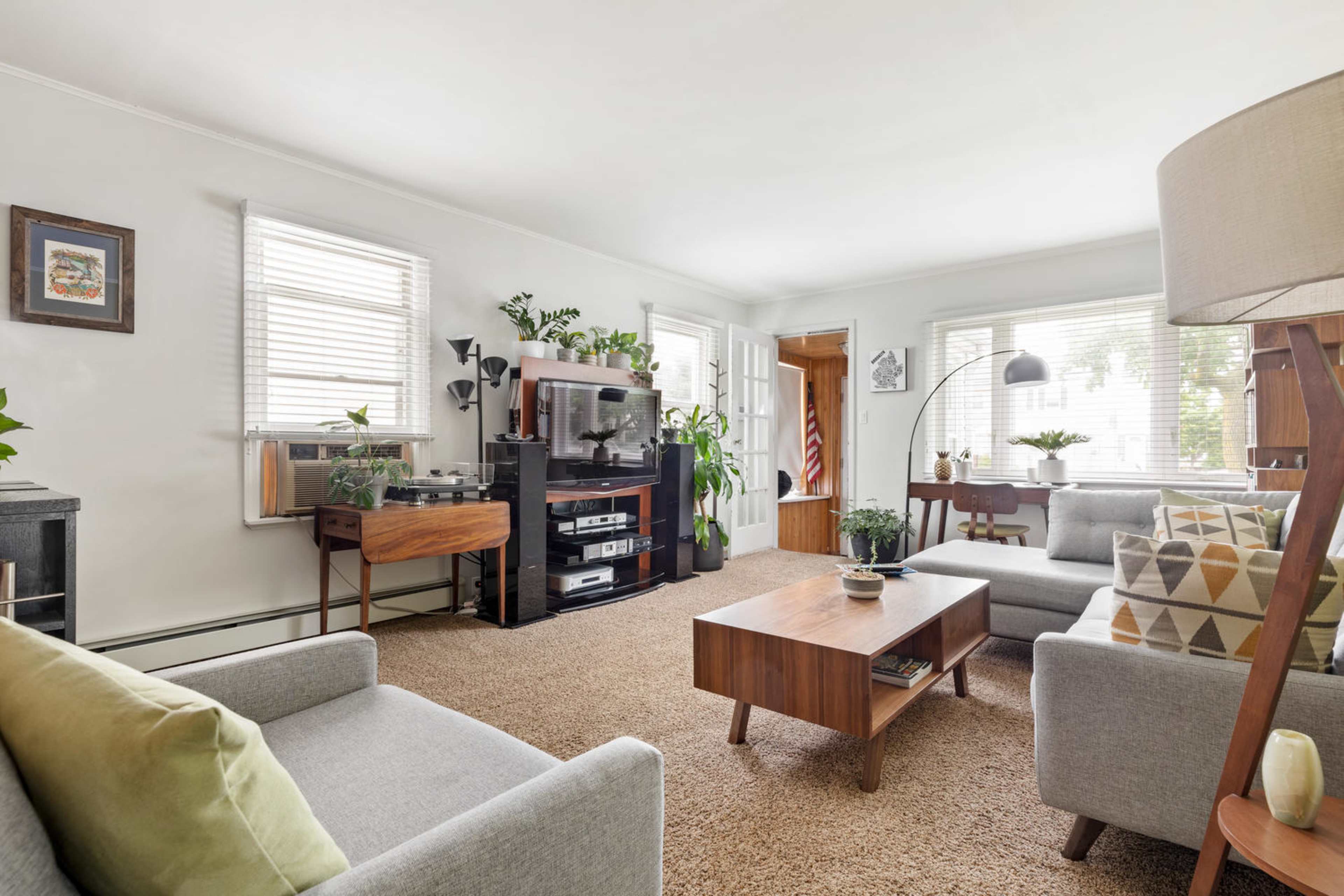 The living room features a comfortable seating arrangement with gray sofas, a wooden coffee table, a media console, and several plants, illuminated by natural light through the windows.