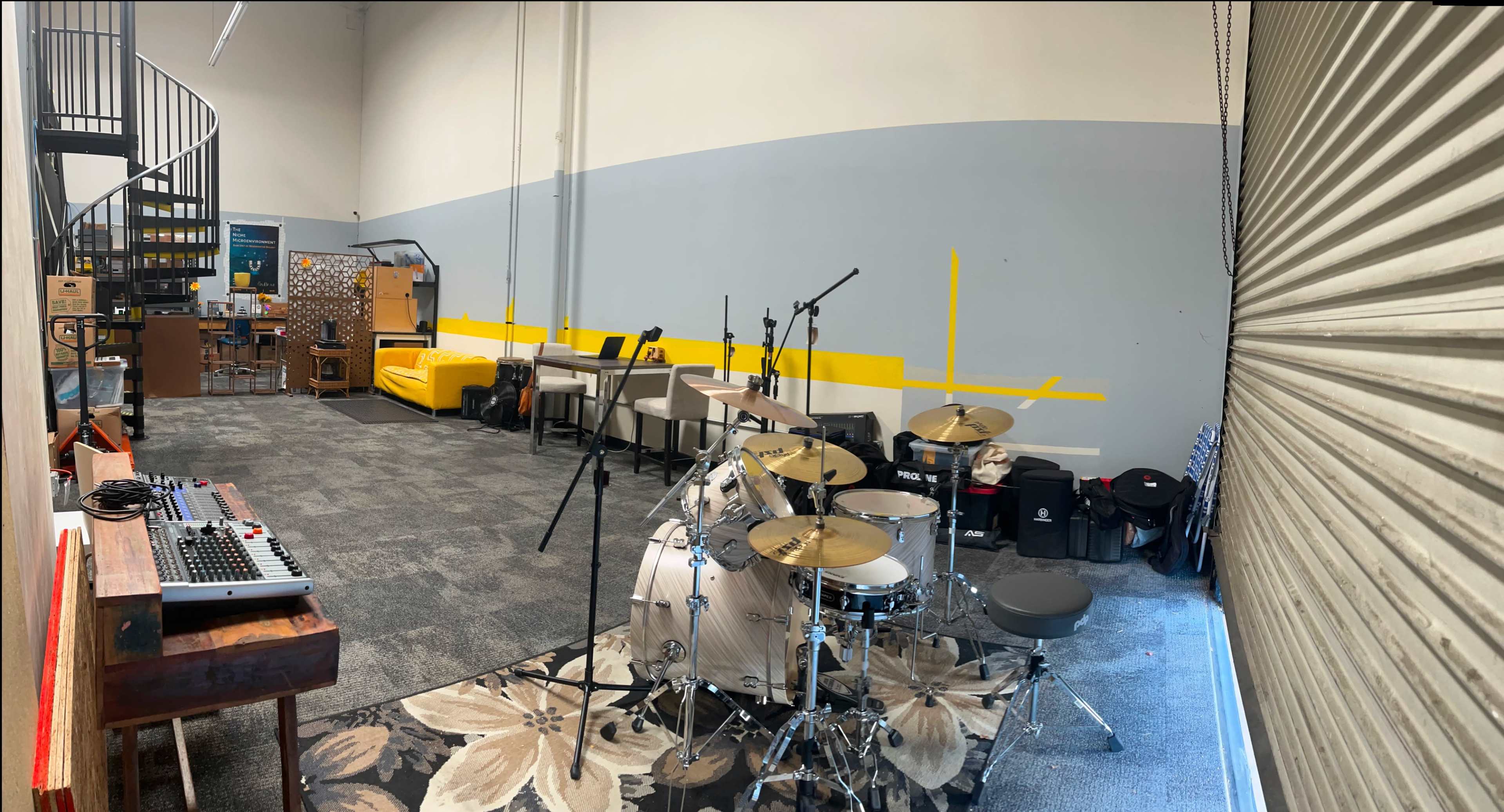 The image shows a spacious indoor room with a drum set and musical equipment on one side, seating areas and a spiral staircase on the left, and yellow accent lines on the gray walls.