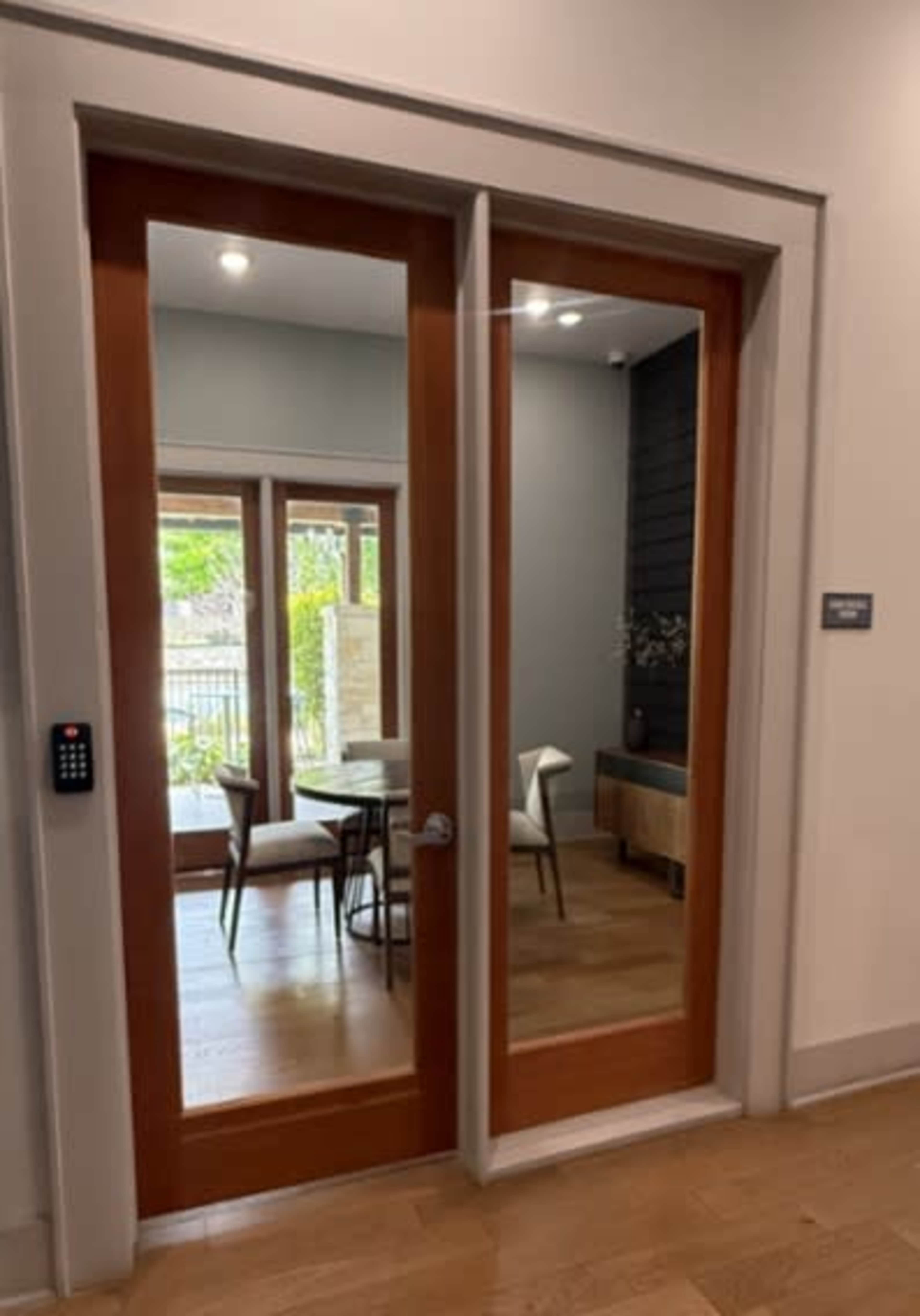 The image shows a pair of glass doors leading to a room with a dining table and chairs, framed by light-colored walls and wooden flooring.