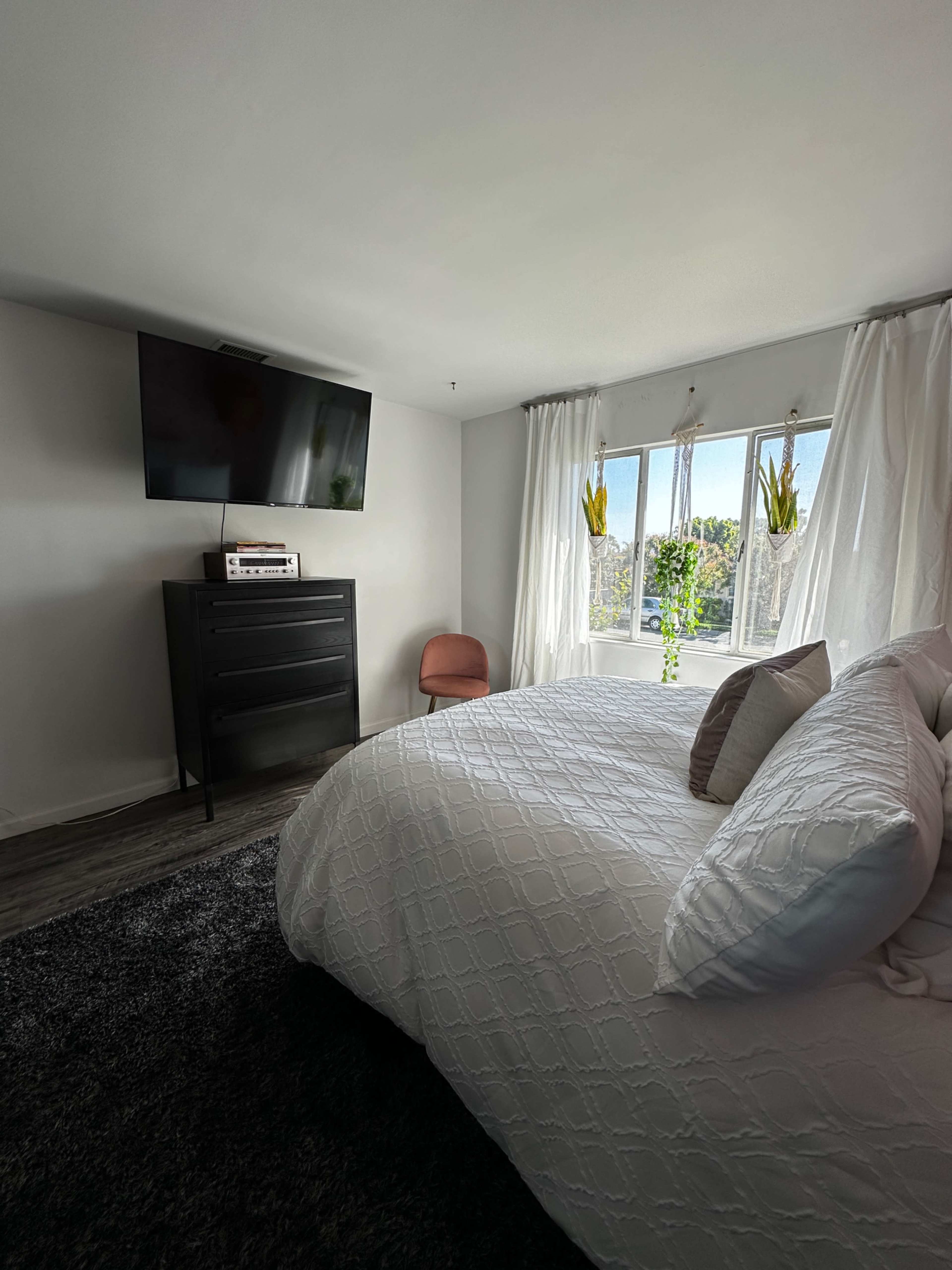 A modern bedroom features a large bed, a dresser, a television mounted on the wall, and bright windows with plants.
