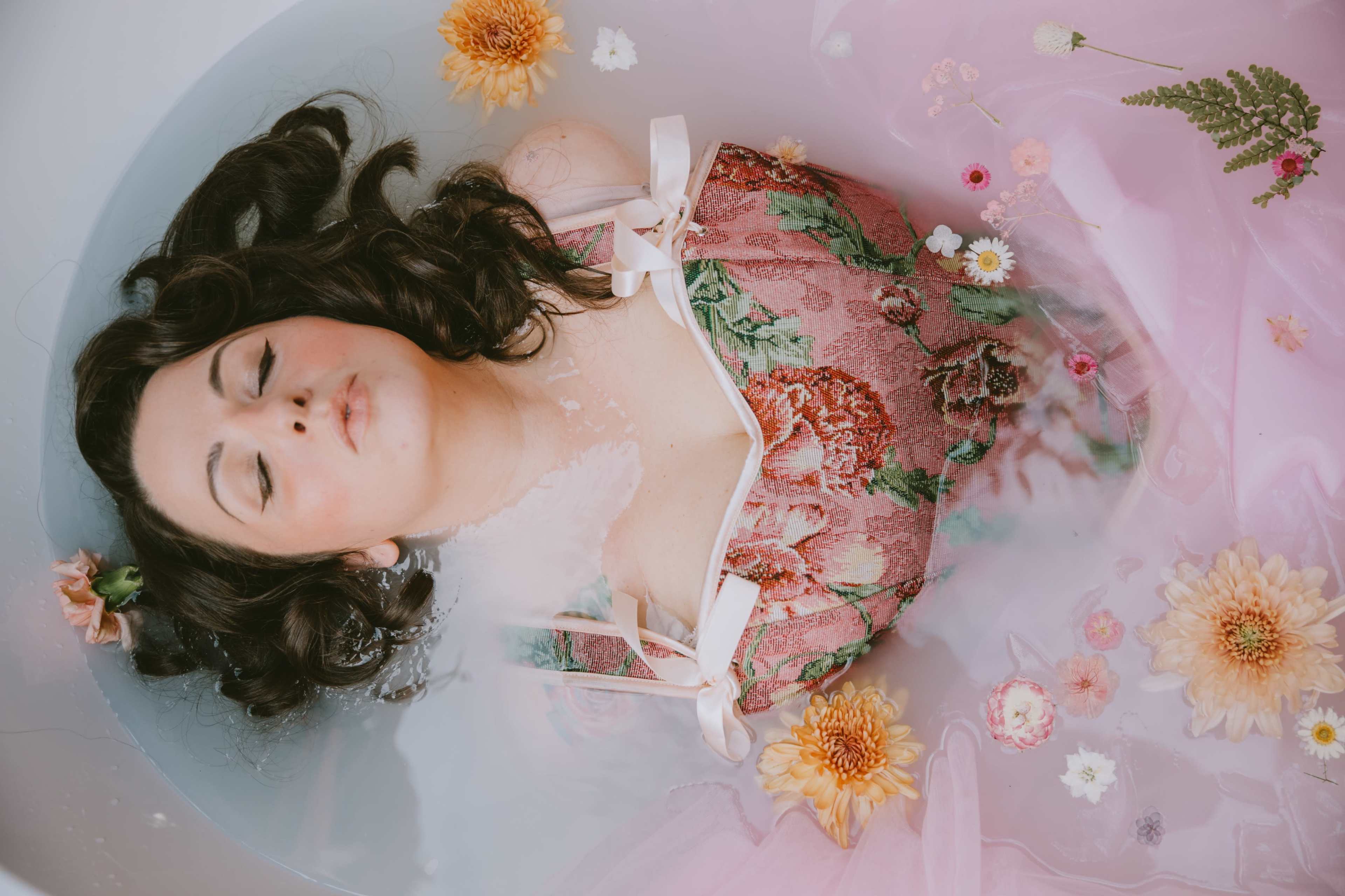 A woman in a floral dress is lying in a bathtub filled with water and floating flowers.