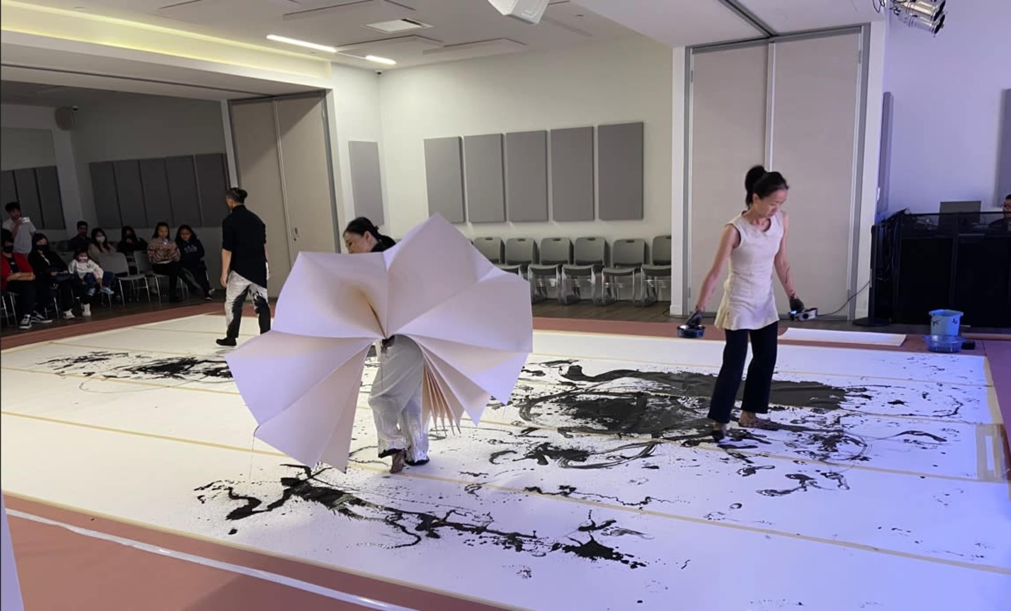 Two artists perform live painting on a large sheet of paper, creating black abstract designs while an audience observes.