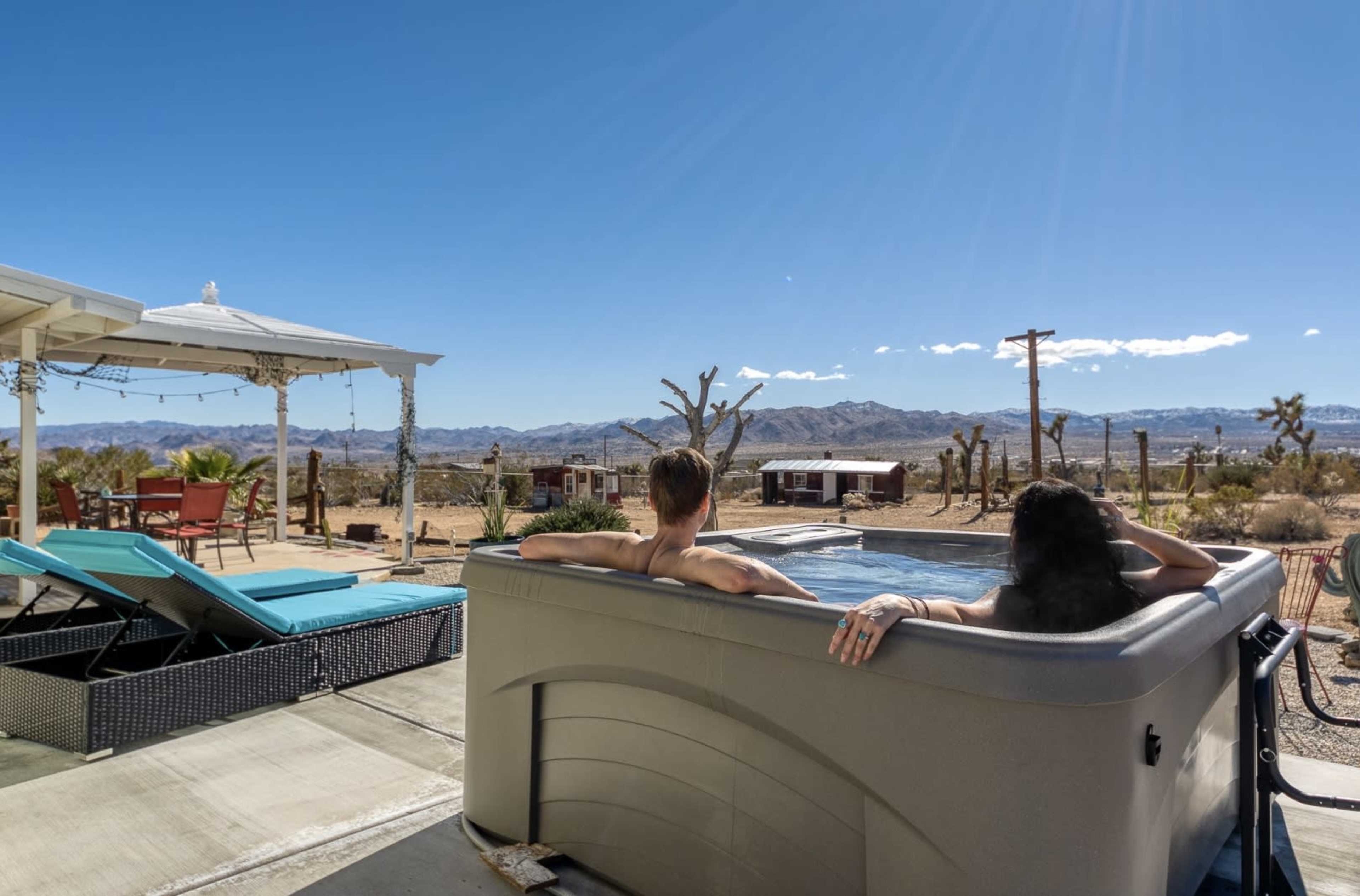 Two people relax in a hot tub overlooking a desert landscape under a clear blue sky.