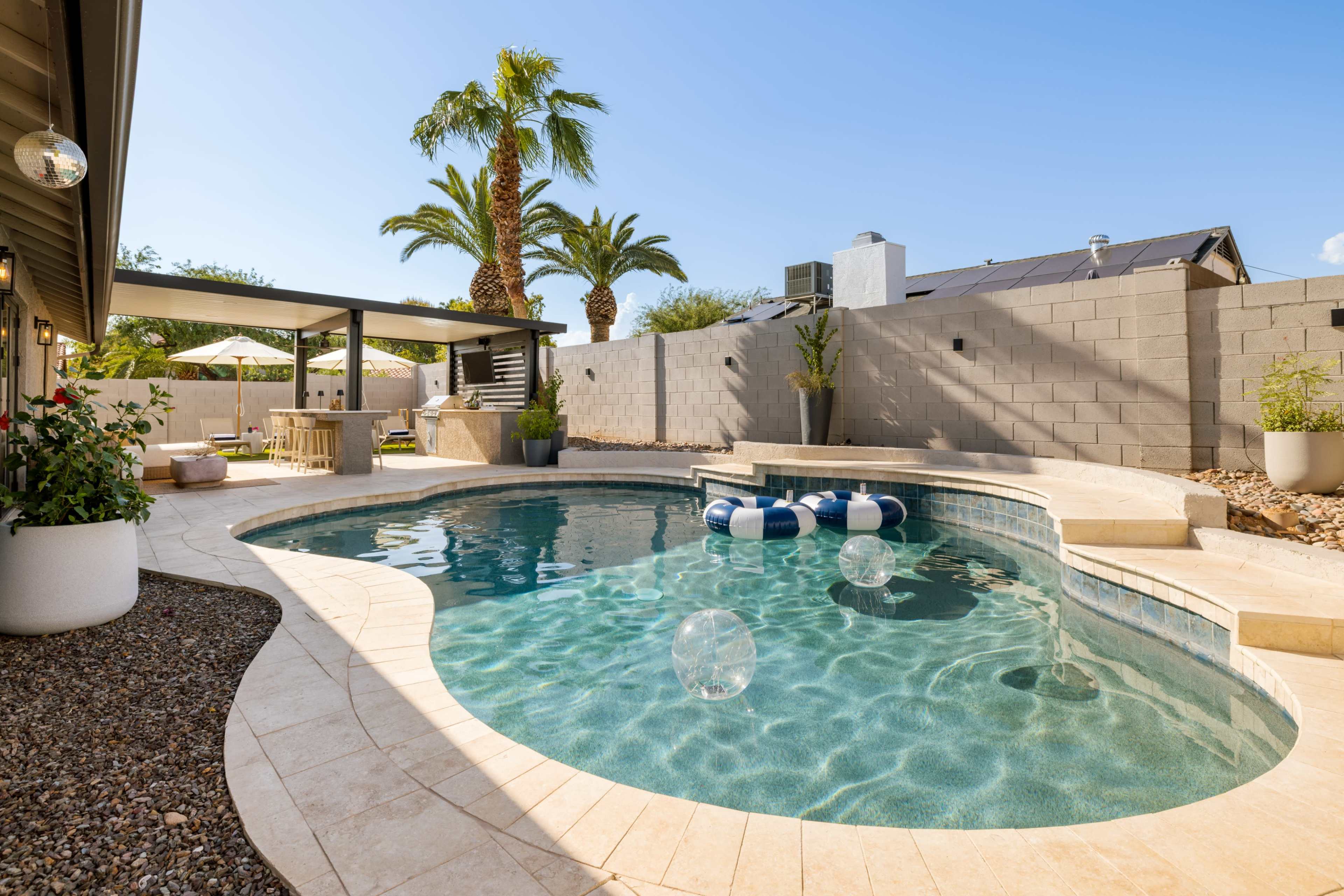 The image shows a backyard with a swimming pool, surrounded by palm trees and a concrete wall, featuring pool floats and a seating area.
