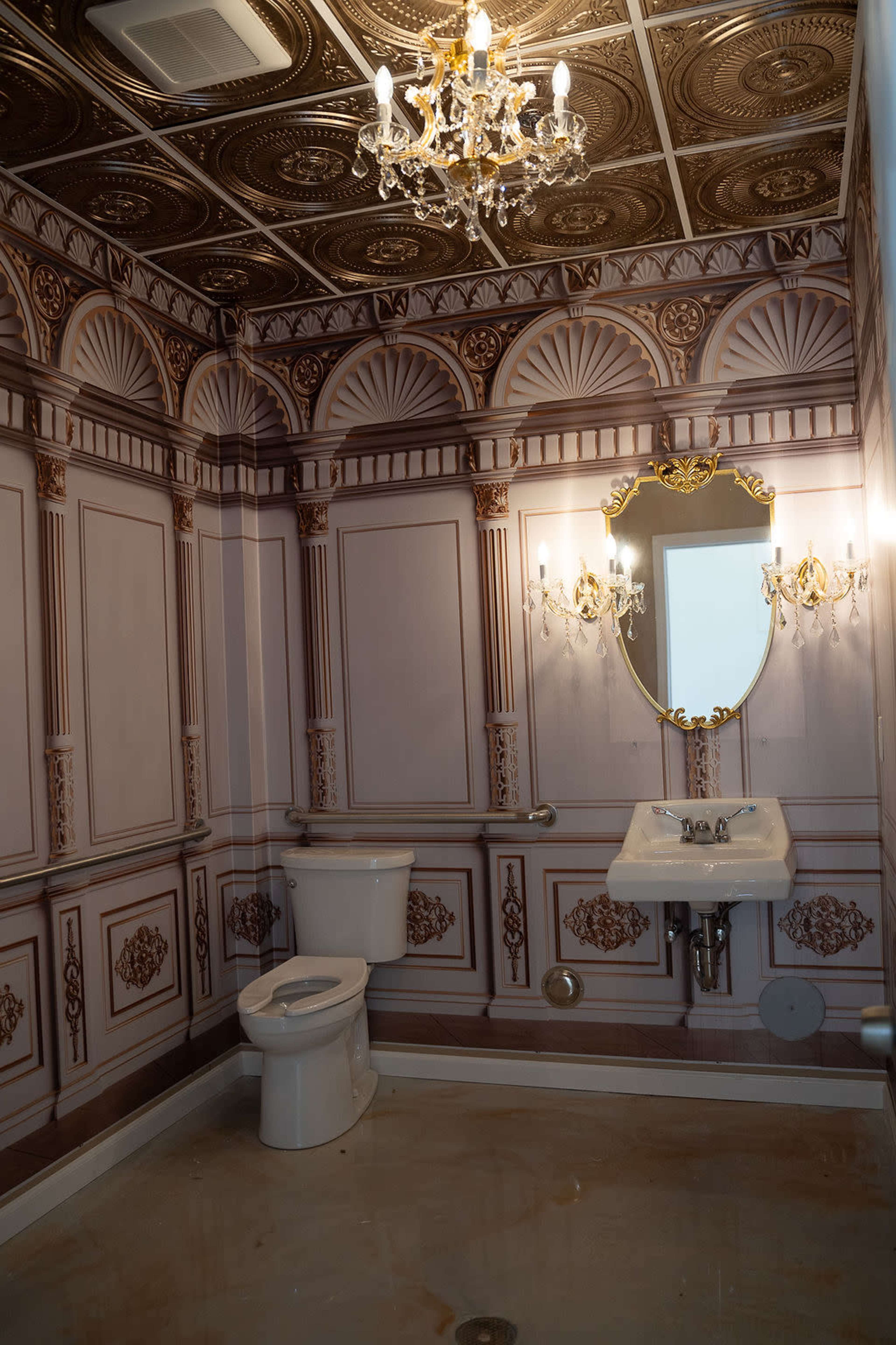 The image shows a small bathroom featuring ornate wallpaper, a chandelier, a wall-mounted sink, and a toilet.
