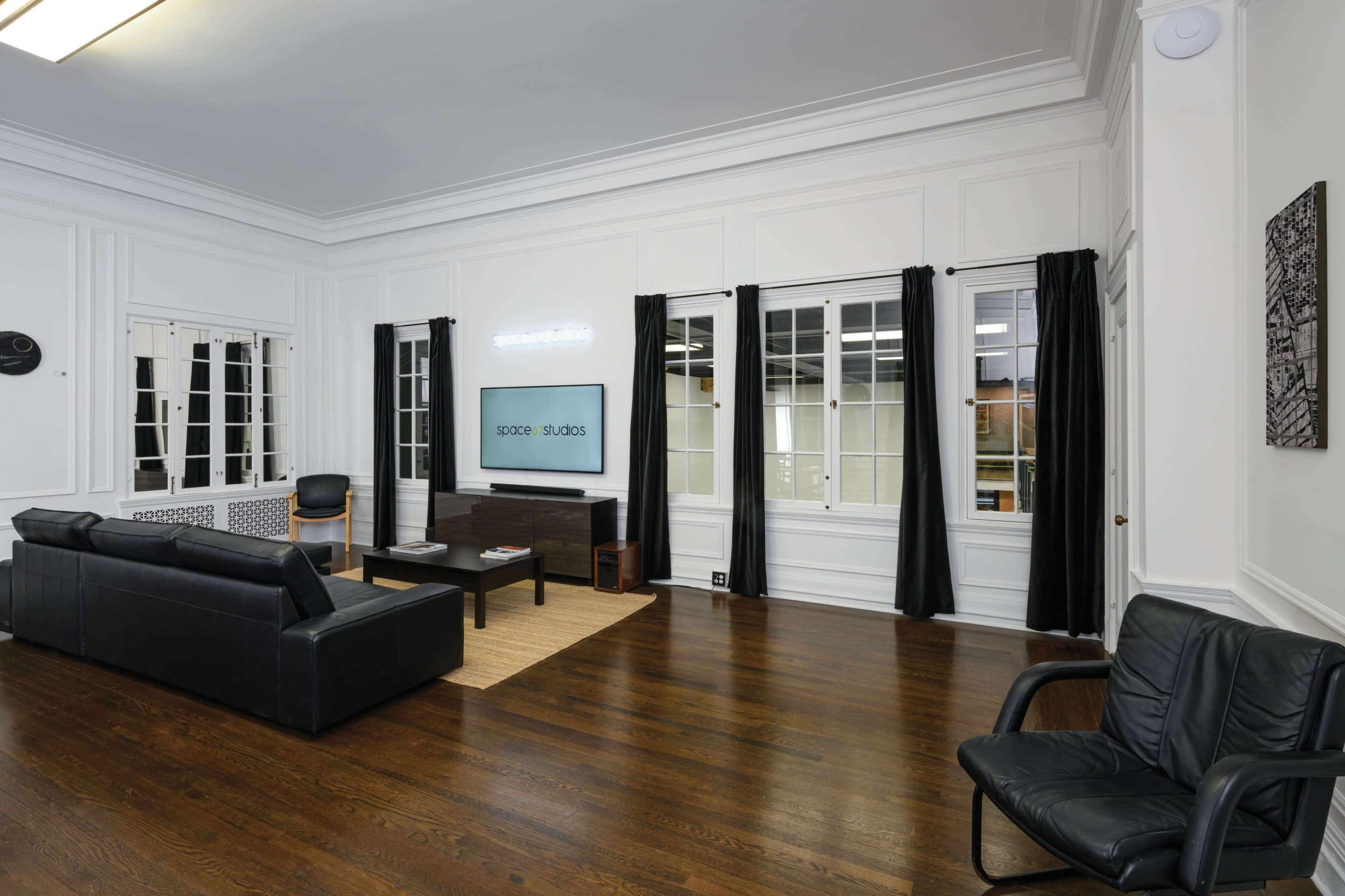 The room features dark hardwood flooring, white walls, large windows with black curtains, and modern furniture including a black couch and chair.