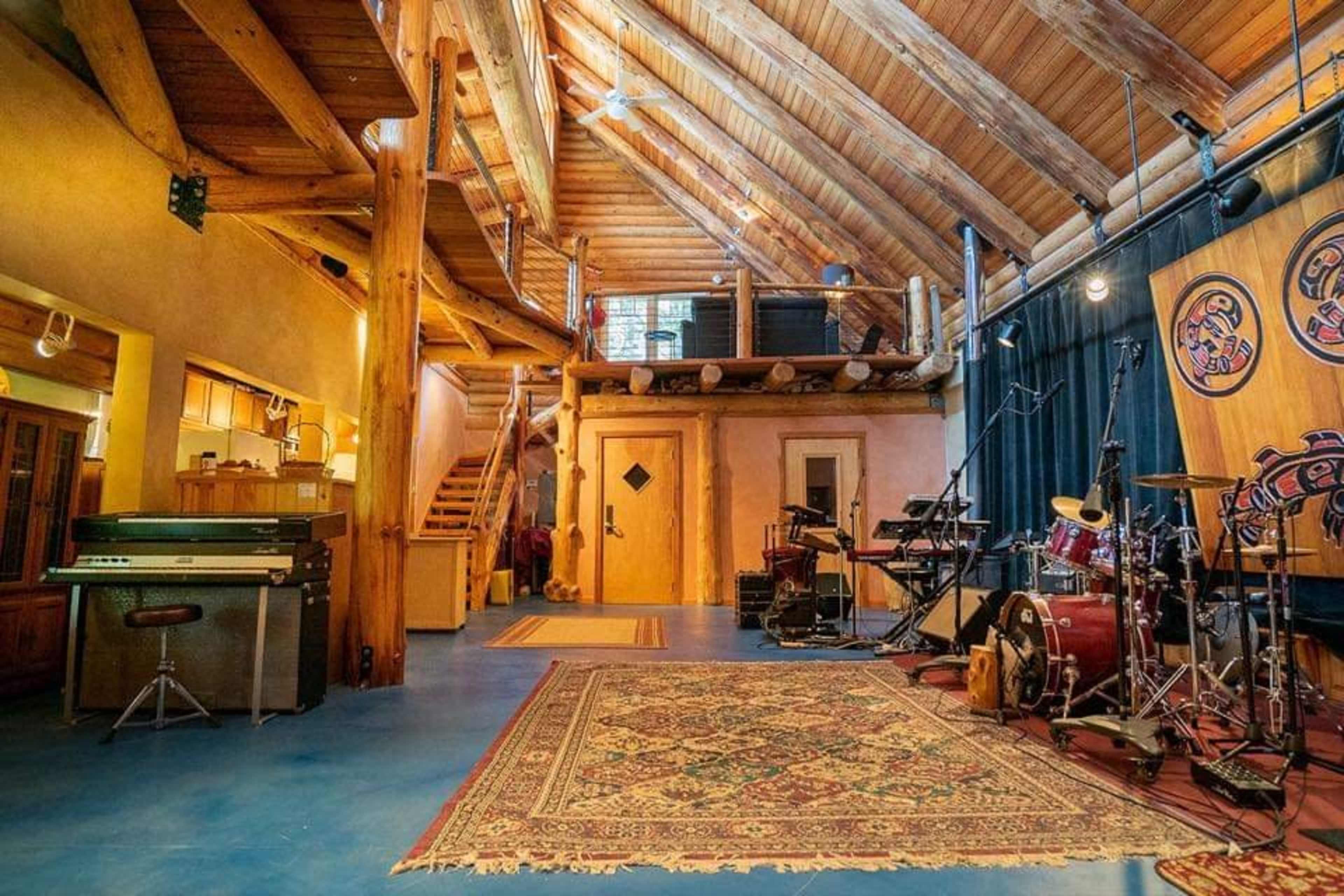 A spacious, wooden interior features a stage with musical equipment, a patterned rug, and a staircase leading to an upper level.